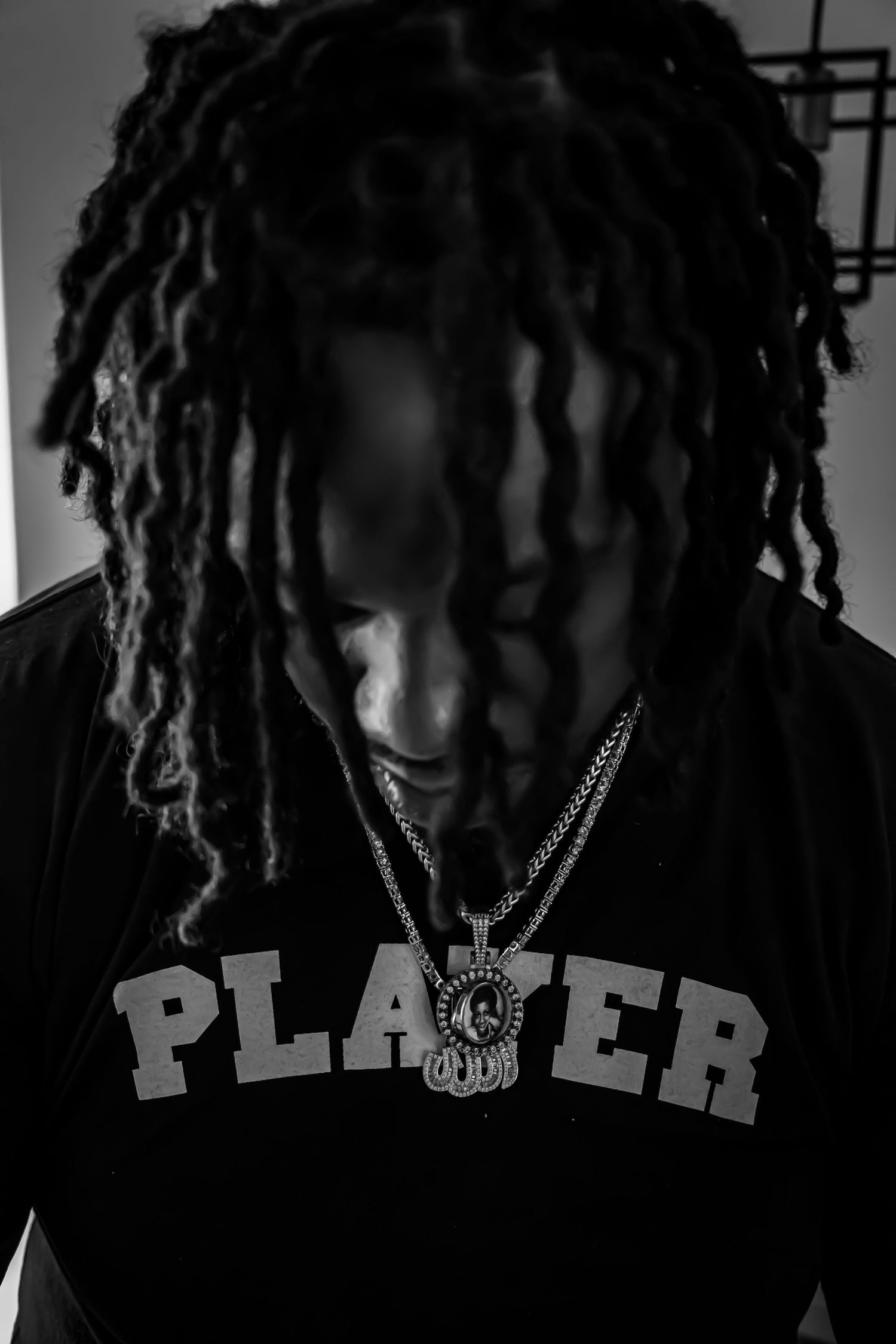 Man with long dreadlocks wearing a black PLAYER t-shirt and chain necklace looking down in moody monochrome portrait.