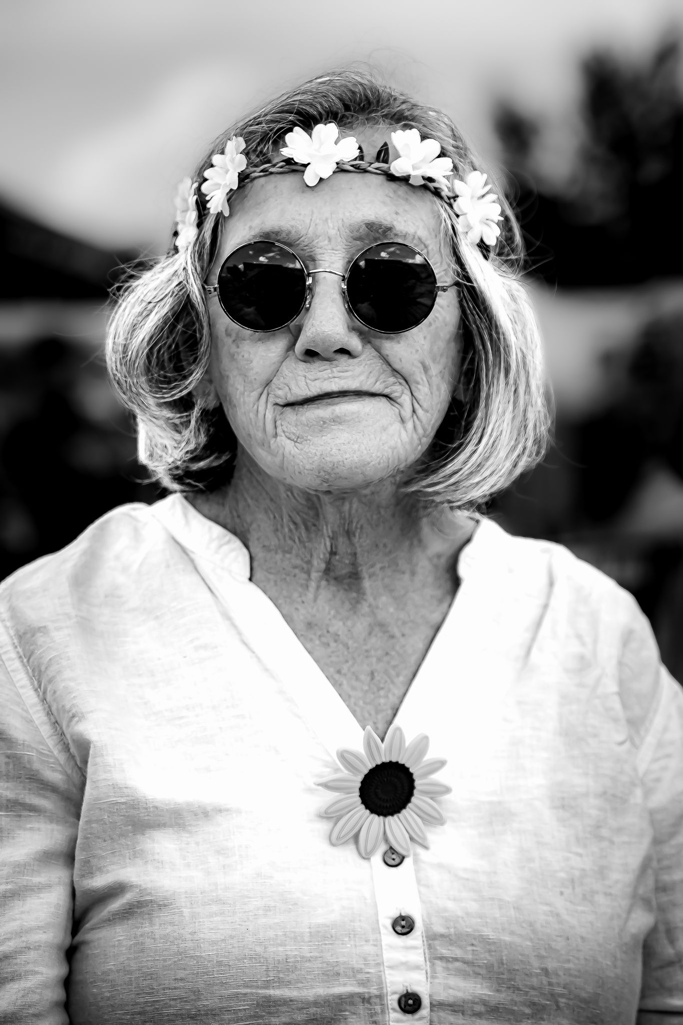Black and white portrait of an elderly woman wearing round sunglasses and a floral crown, smiling softly outdoors.