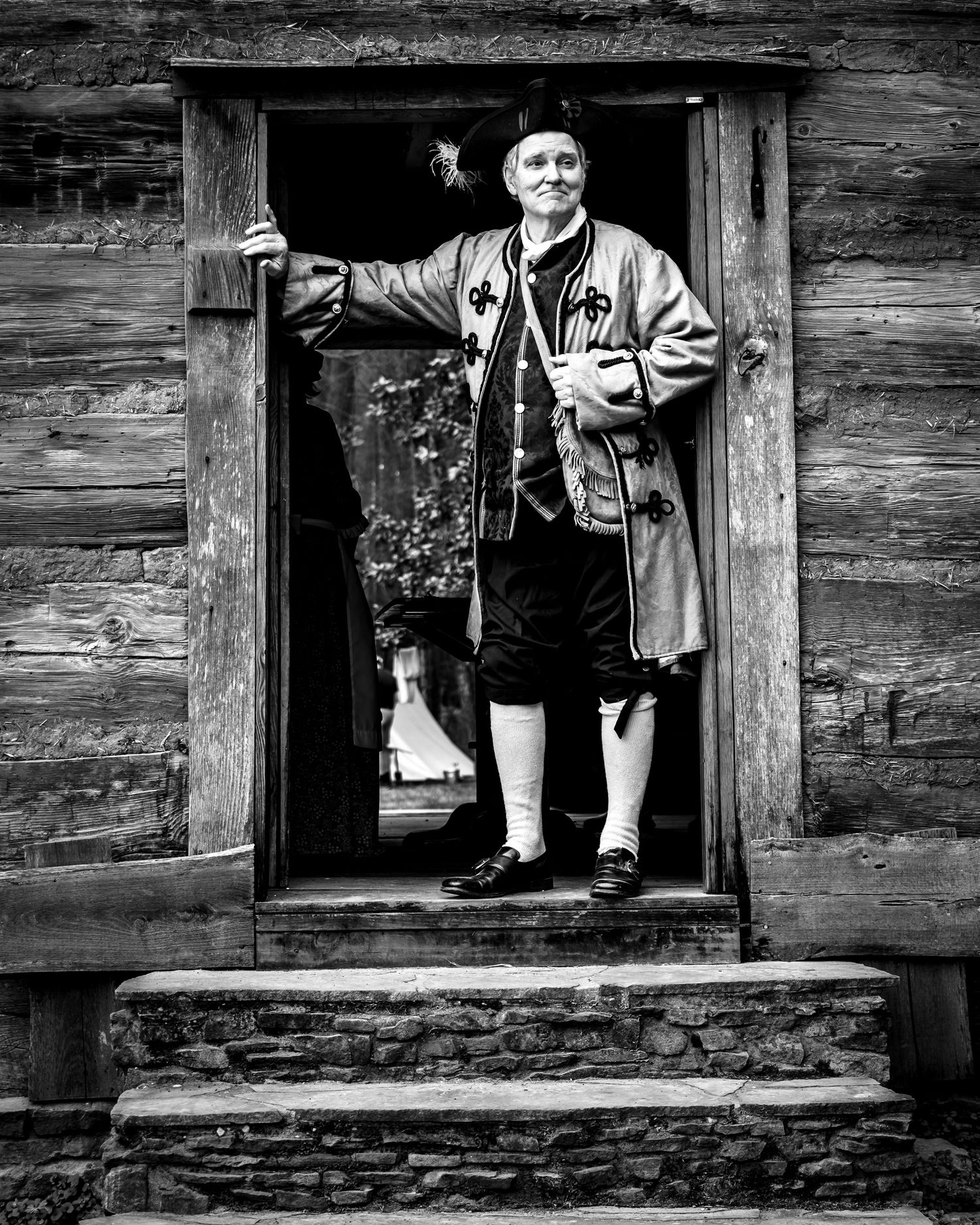 Man in 18th century colonial costume stands in doorway of rustic log building, black and white photography.