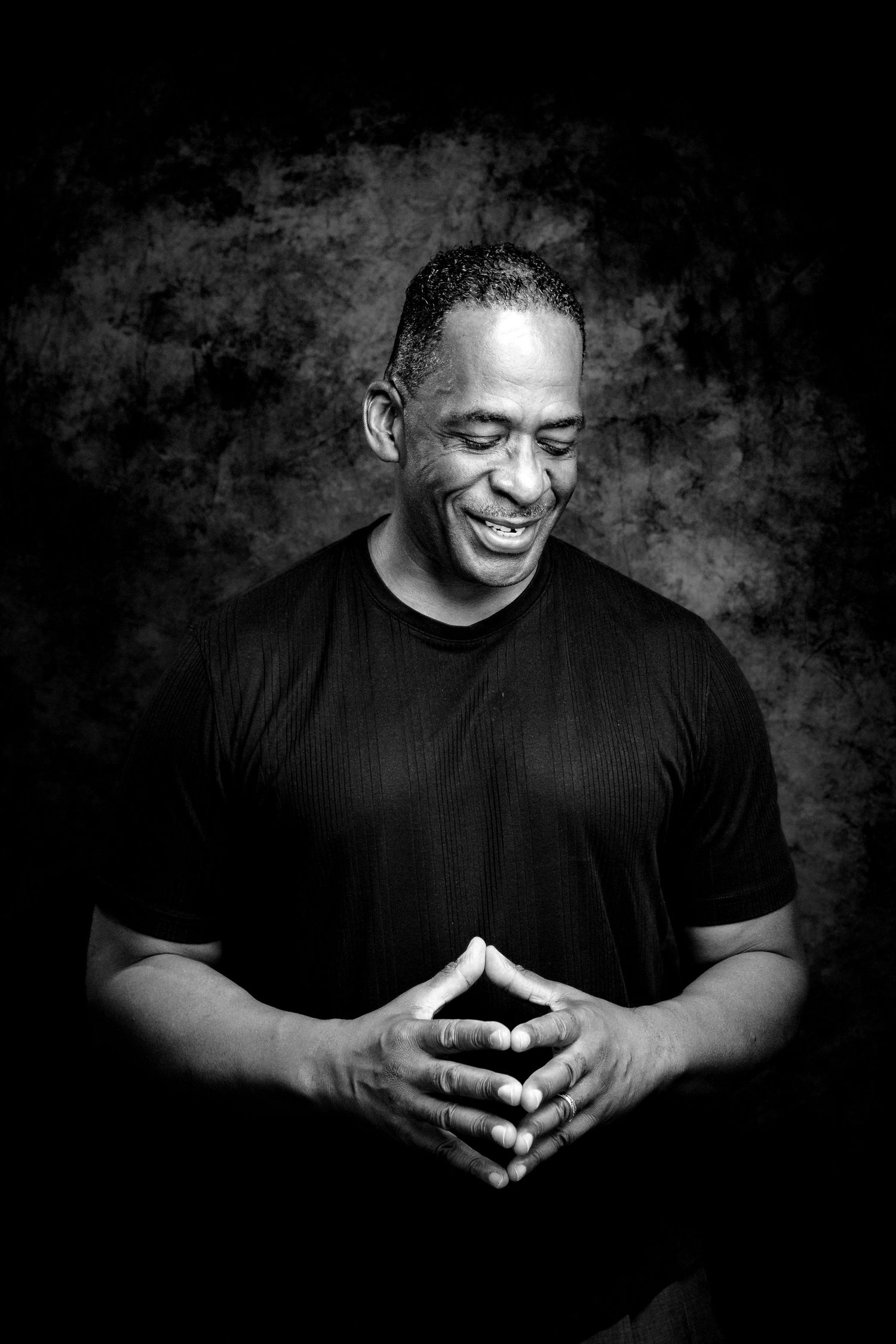 Black and white portrait of a smiling man in a black t-shirt with clasped hands against dark background.