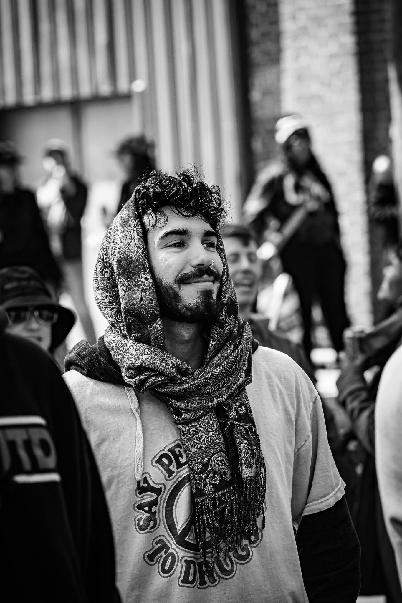 Black and white portrait of a smiling bearded man wearing a peace sign t-shirt and patterned scarf on his head.