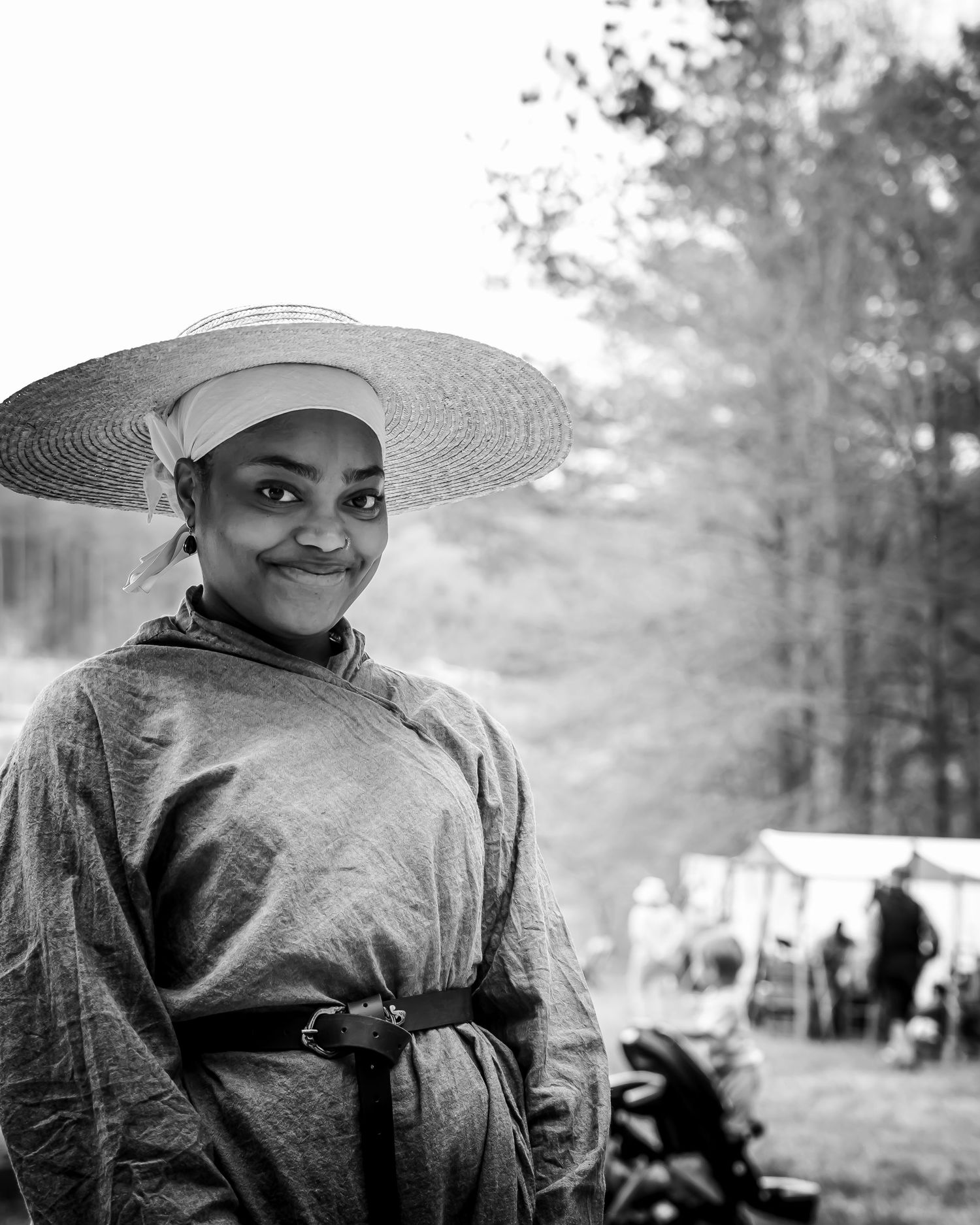 Smiling woman in period costume and straw hat at outdoor historical reenactment, black and white photo.