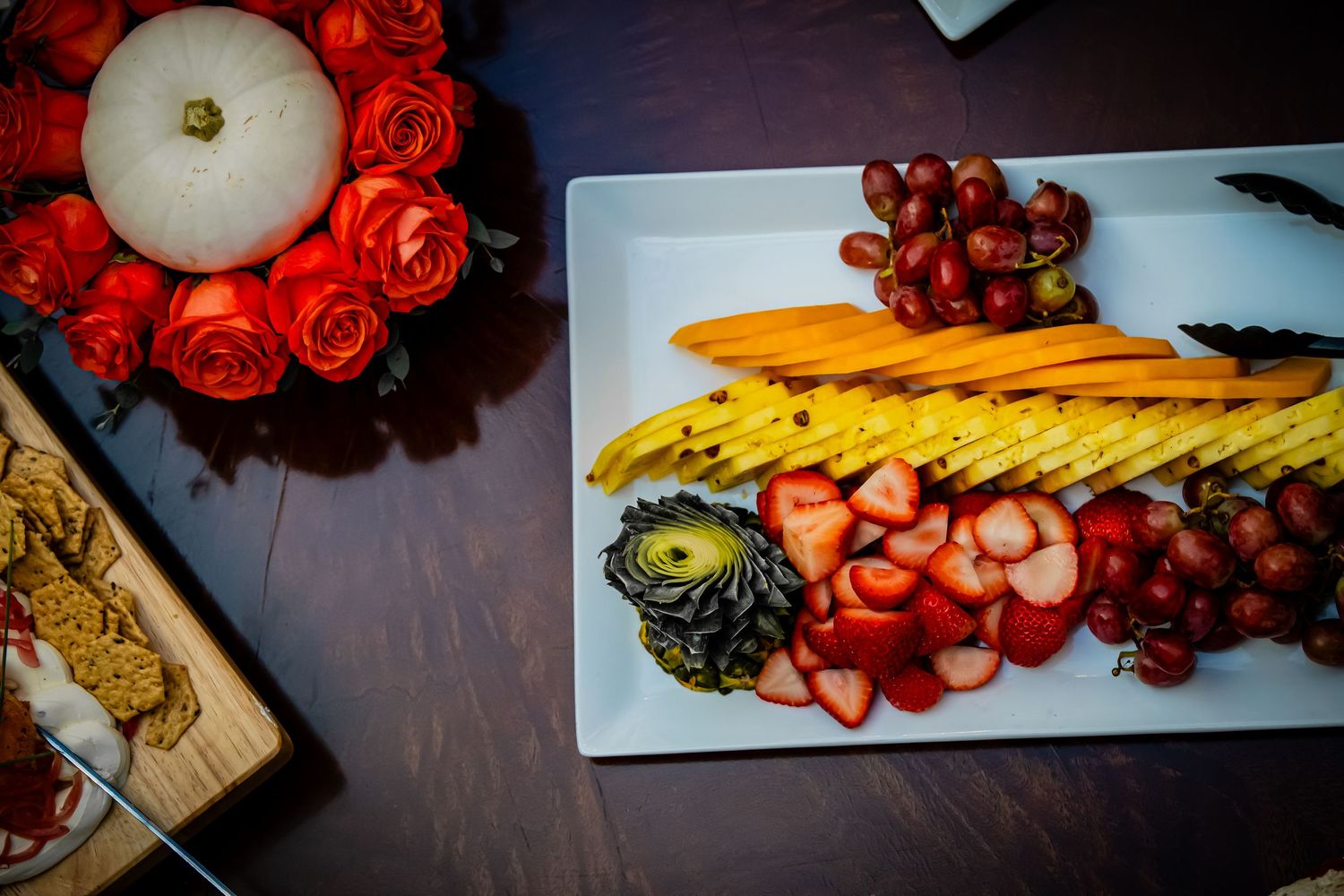 Overhead view of a colorful fruit platter with strawberries, grapes, and mango slices beside a floral centerpiece.