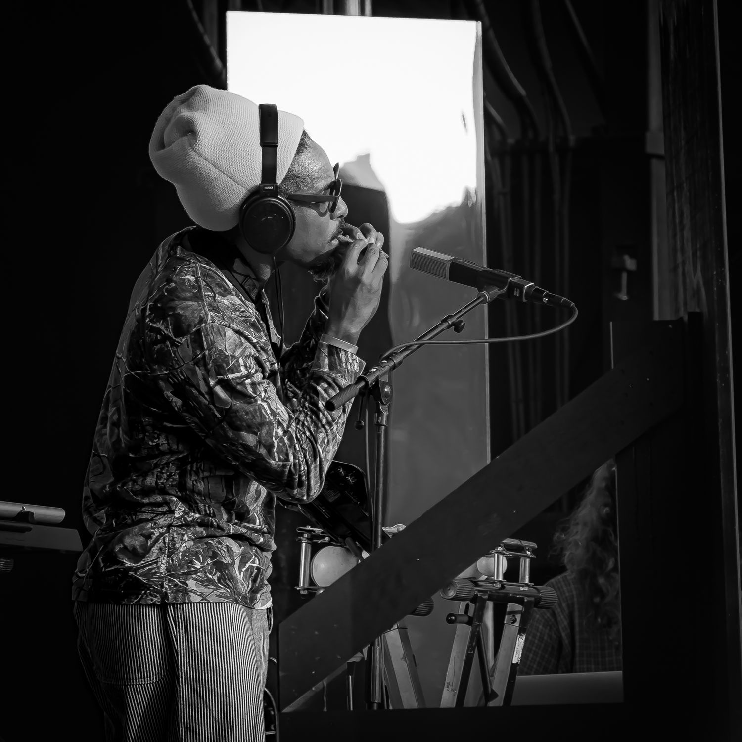 Black and white photo of a musician wearing headphones and a hat singing into a microphone on stage.