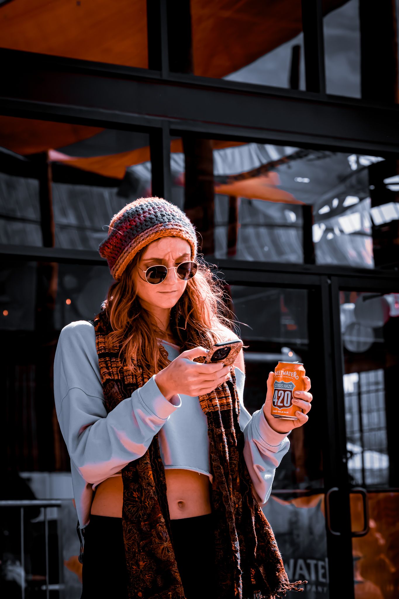 Color photo of a young woman in a beanie hat and sunglasses holding an orange beverage can outdoors.