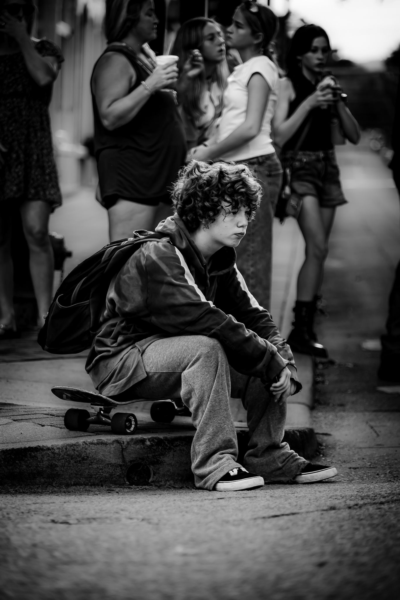 Black and white photo of a teenage boy sitting on a skateboard outdoors, wearing a hoodie and backpack.
