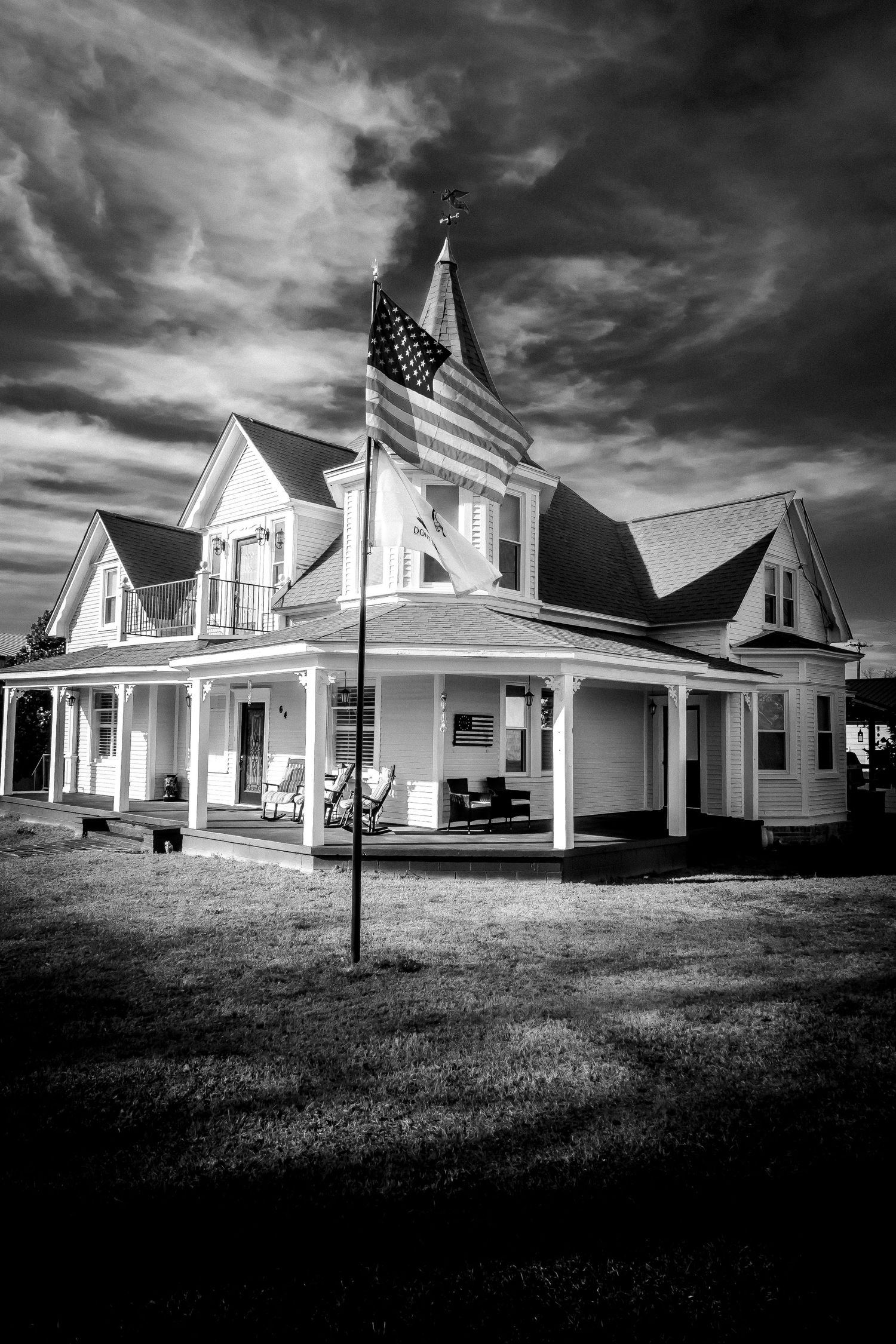Black and white photo of a Victorian-style white house with wrap-around porch and American flag.