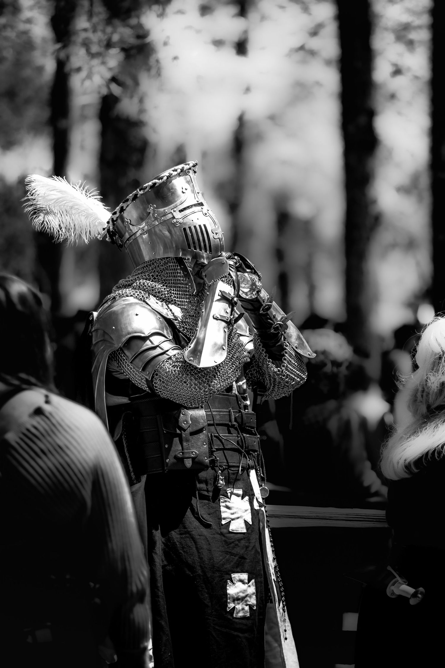 Knight in full plate armor with feathered helmet stands in a forest in black and white photo.