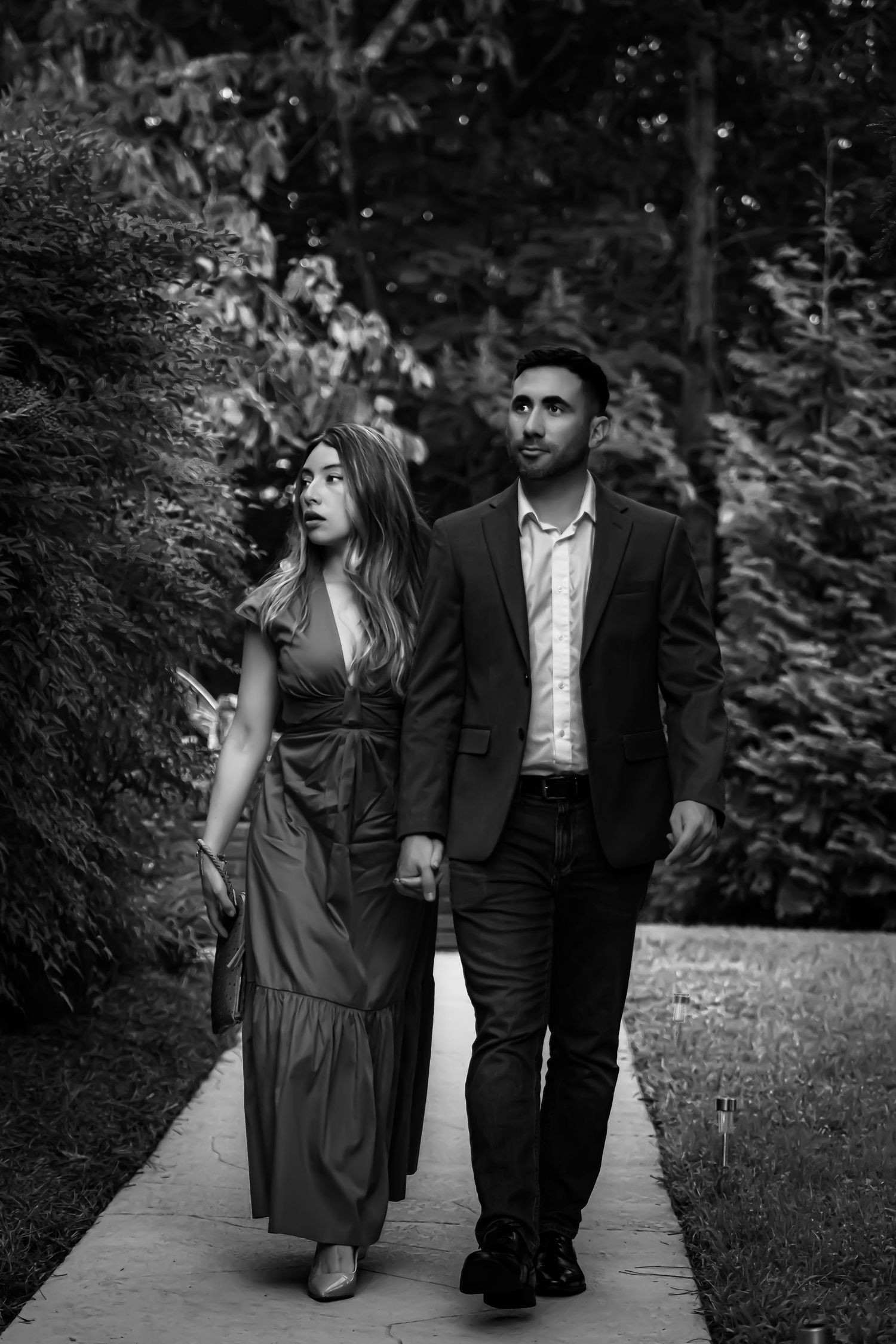 Couple holding hands walking along a garden path in a black and white outdoor portrait.