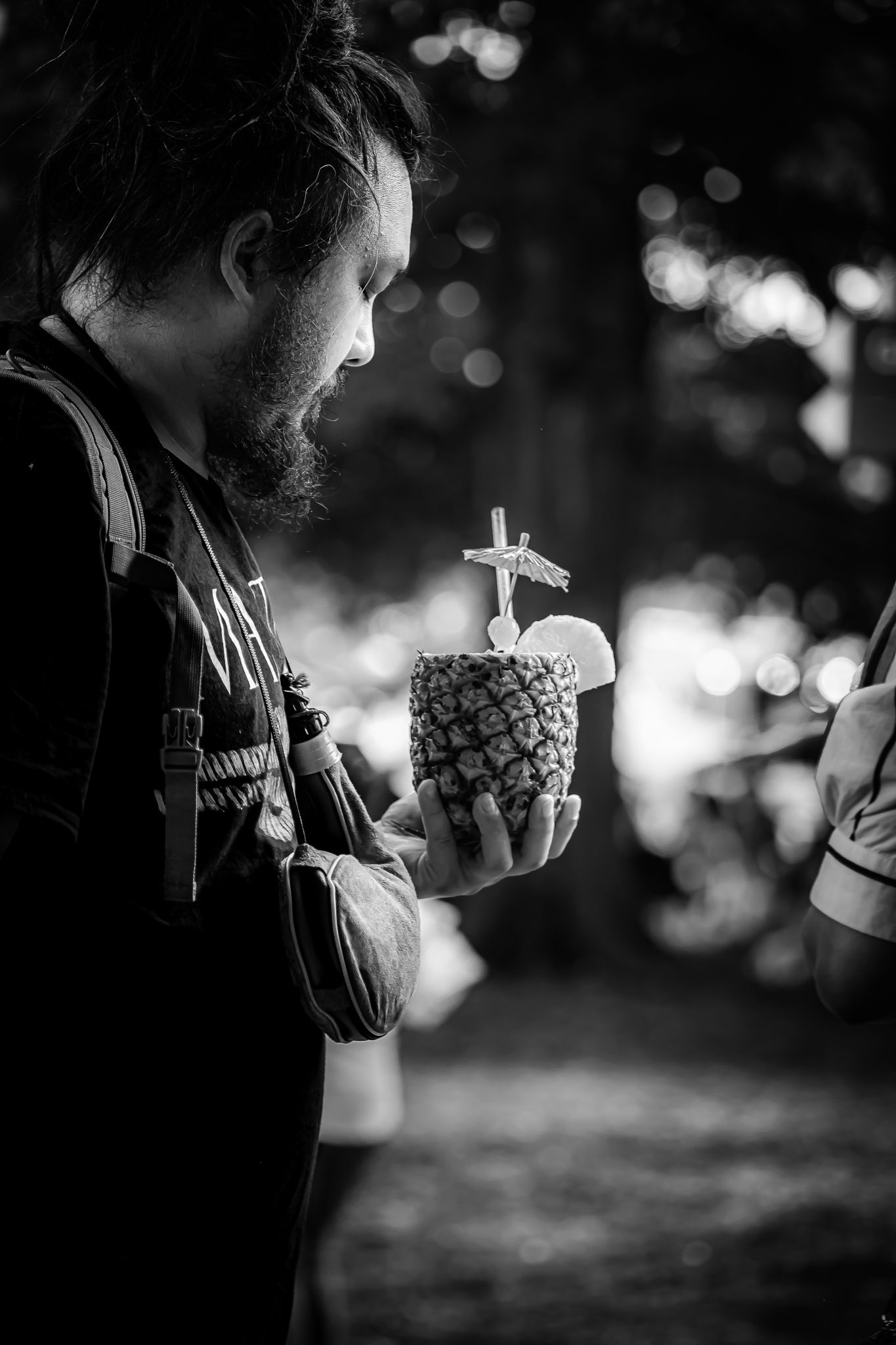 Bearded man with dreadlocks examines a pineapple cocktail with straw in moody black and white bokeh photo.