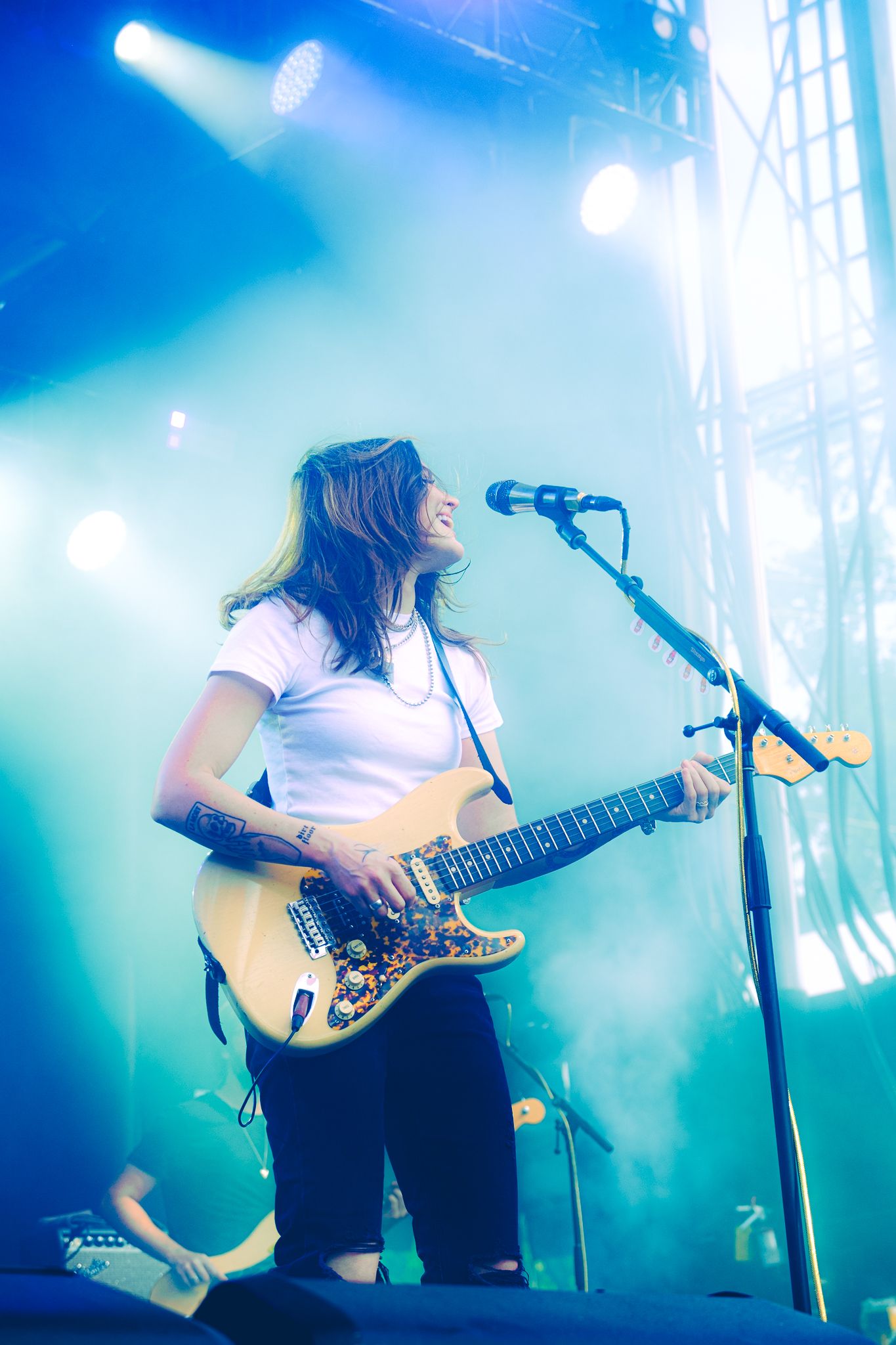 Female guitarist smiling while playing Fender Stratocaster on stage surrounded by blue stage lighting.