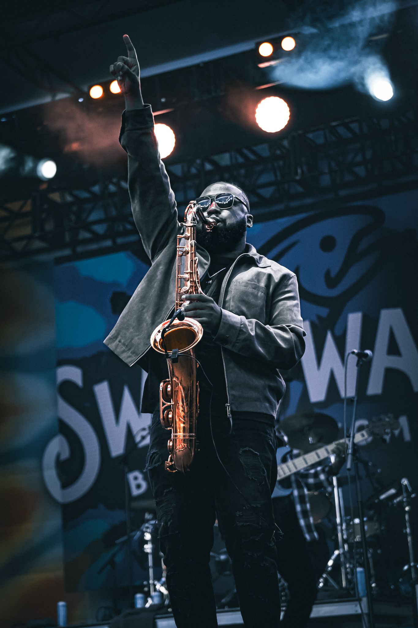 Saxophonist raises fist triumphantly on stage under bright lights at an outdoor music festival concert.