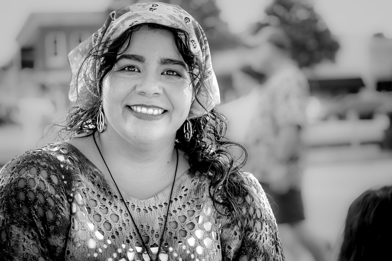 Smiling woman wearing a patterned headscarf and lace top with a necklace in a black and white portrait photo.