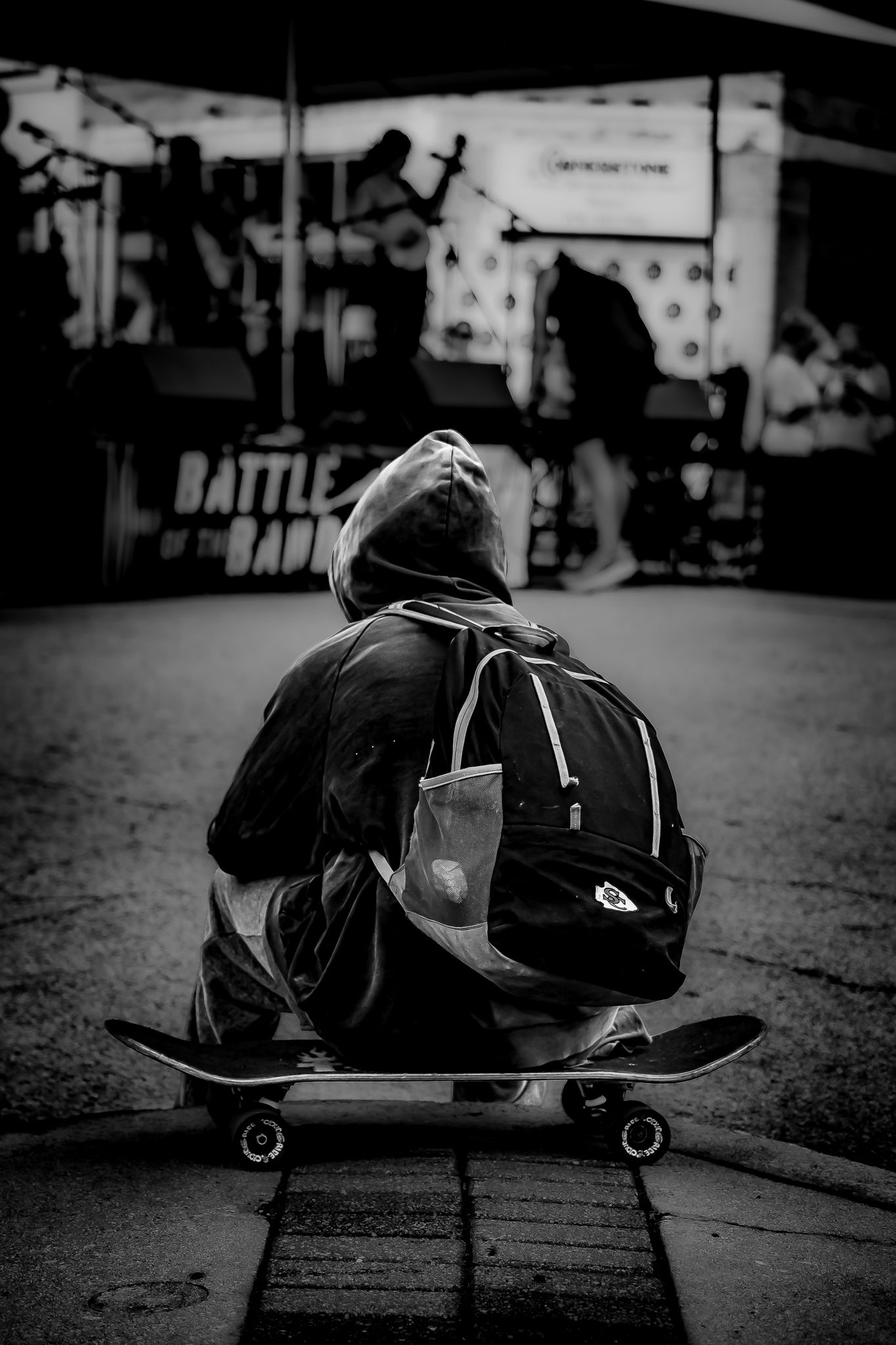 Black and white photo of a person sitting on a skateboard wearing a hoodie and backpack, watching a Battle of the Bands event.