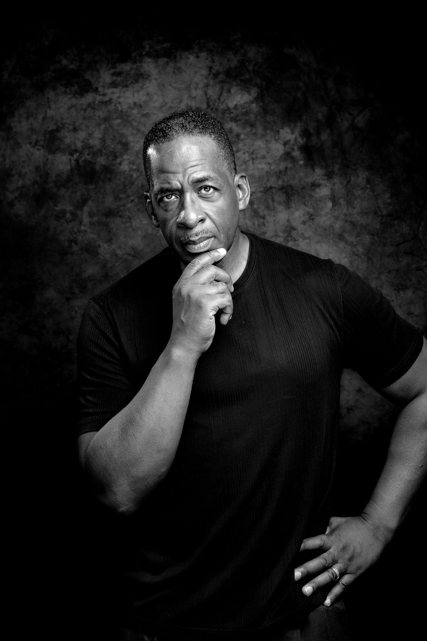 Black and white studio portrait of a thoughtful middle-aged man in black t-shirt with hand on chin.