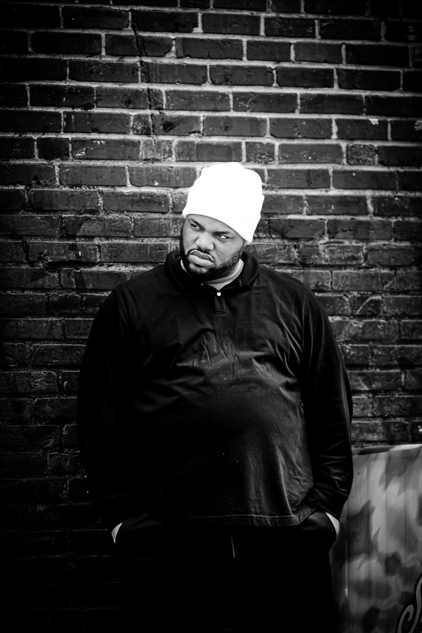 Black and white portrait of a man in a black jacket and white beanie leaning against a brick wall.