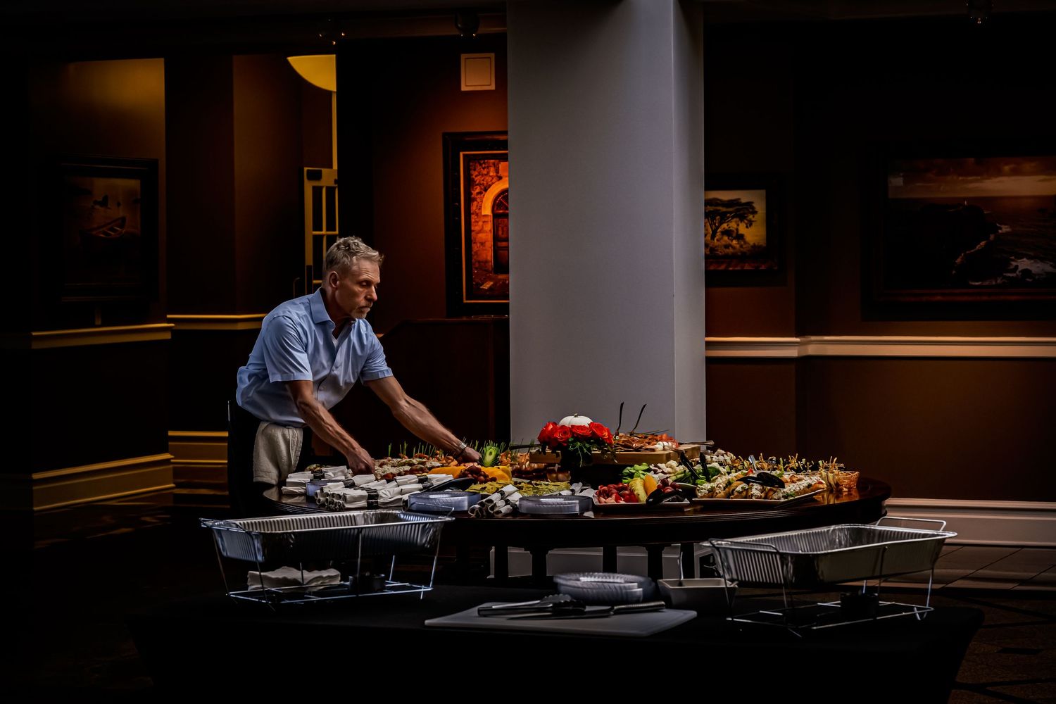 Catering professional arranging an elaborate food display on a banquet table in a dimly lit event hall.