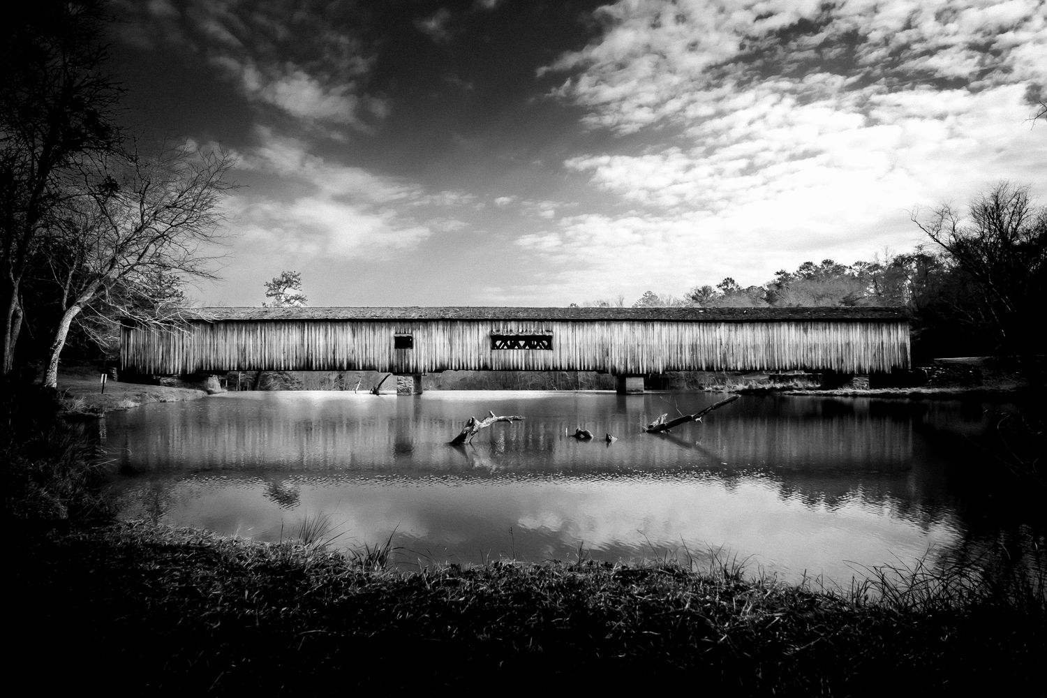 Black and white photo of a historic covered bridge reflected in calm water surrounded by bare trees.