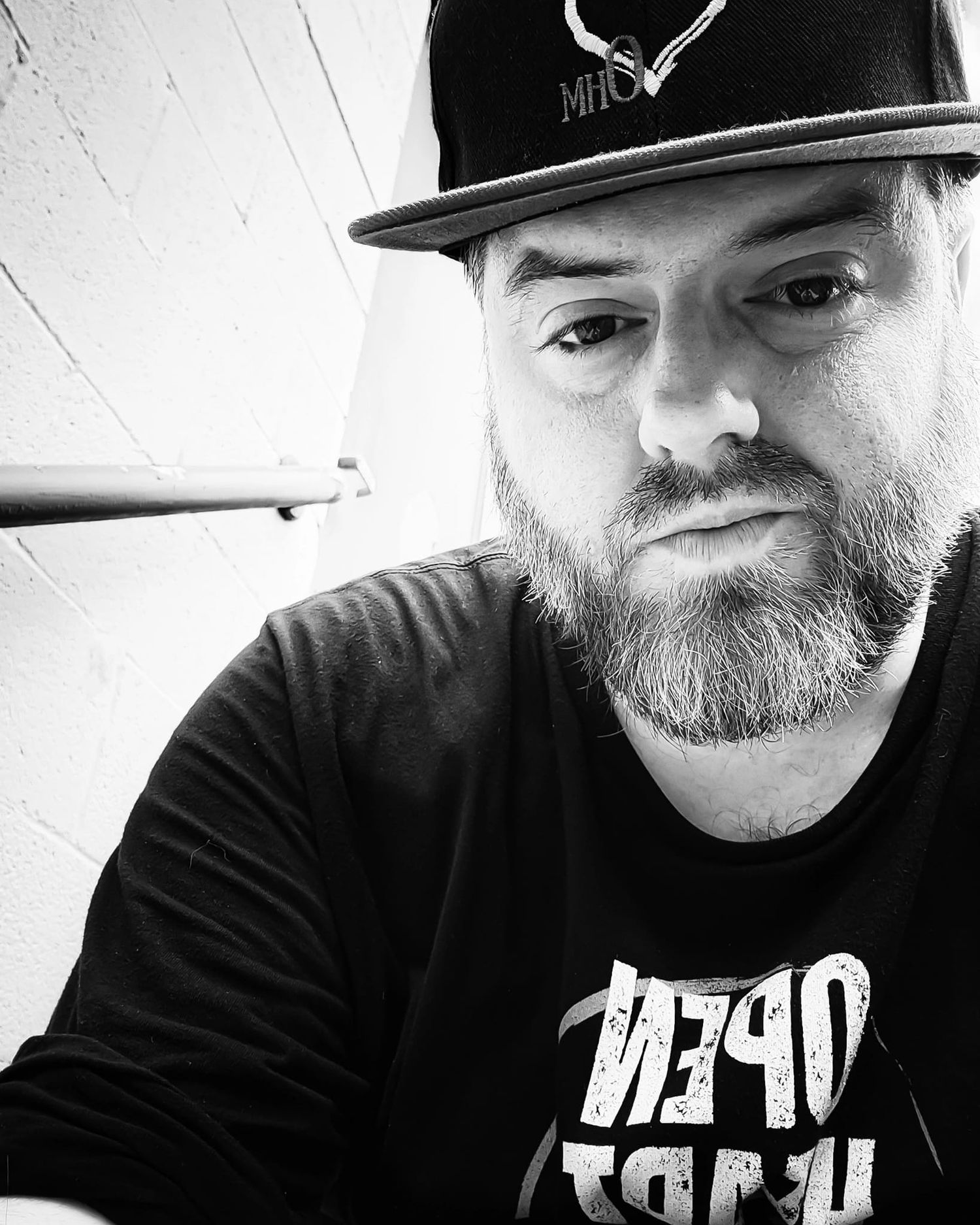 Black and white close-up portrait of a bearded man wearing a snapback cap and black graphic t-shirt.