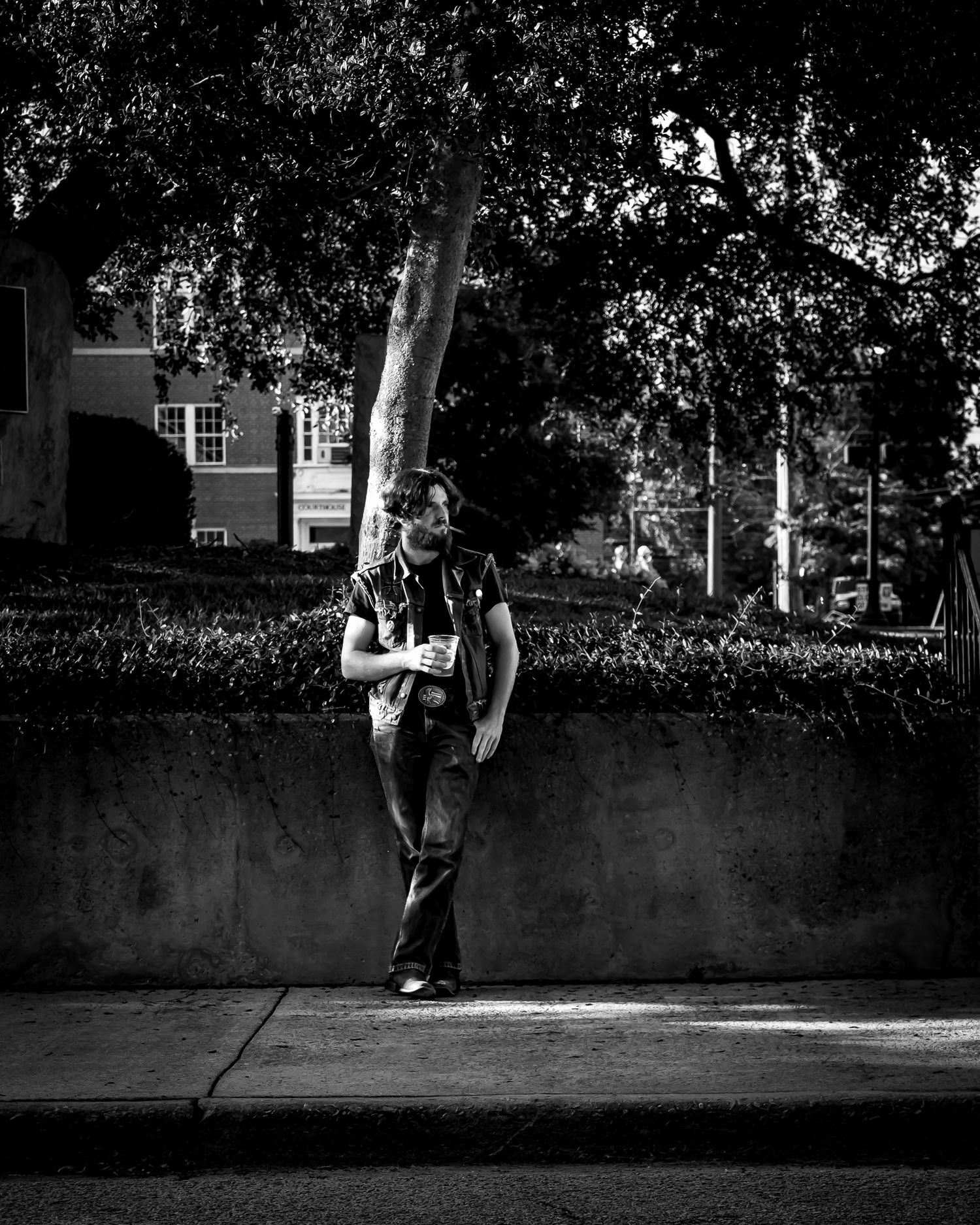 Black and white photo of a person standing on a sidewalk beside a concrete wall, trees and building in background.