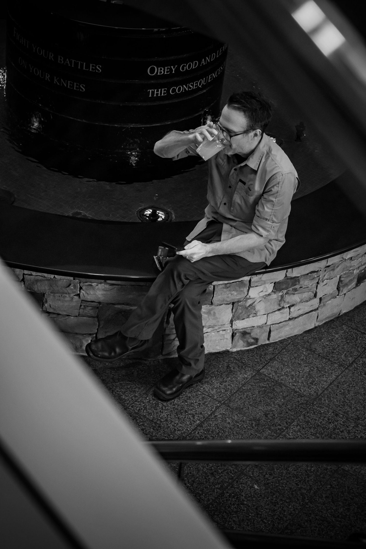 Black and white photo of a man sitting on a stone ledge drinking from a cup in a dimly lit venue.