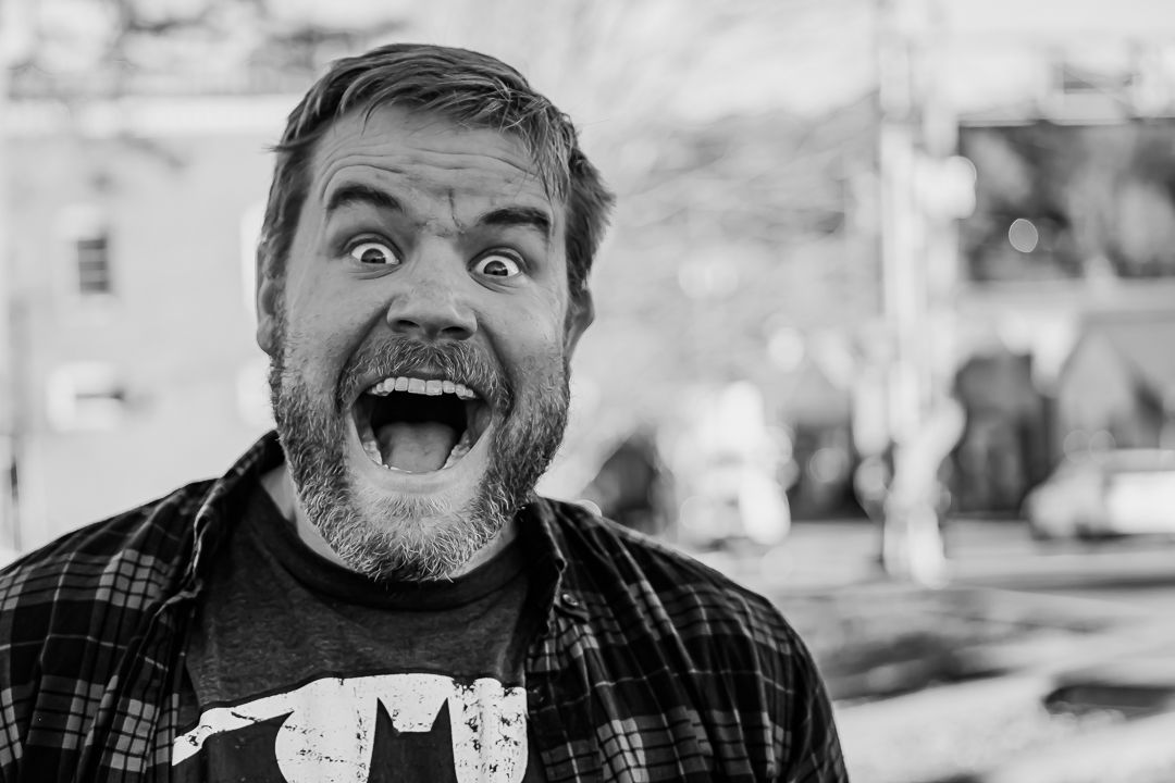 Black and white close-up of a bearded man with wide eyes and open mouth making a surprised expression outdoors.