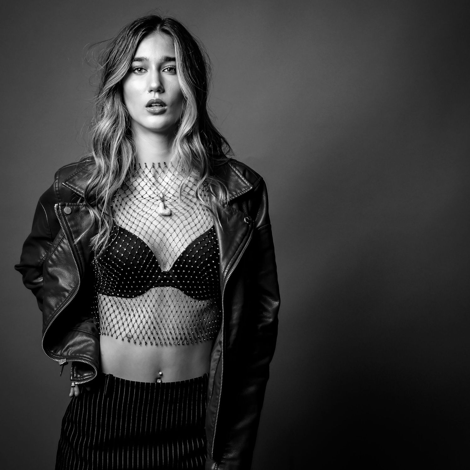 Woman in leather jacket and mesh crop top poses confidently in black and white studio photo.