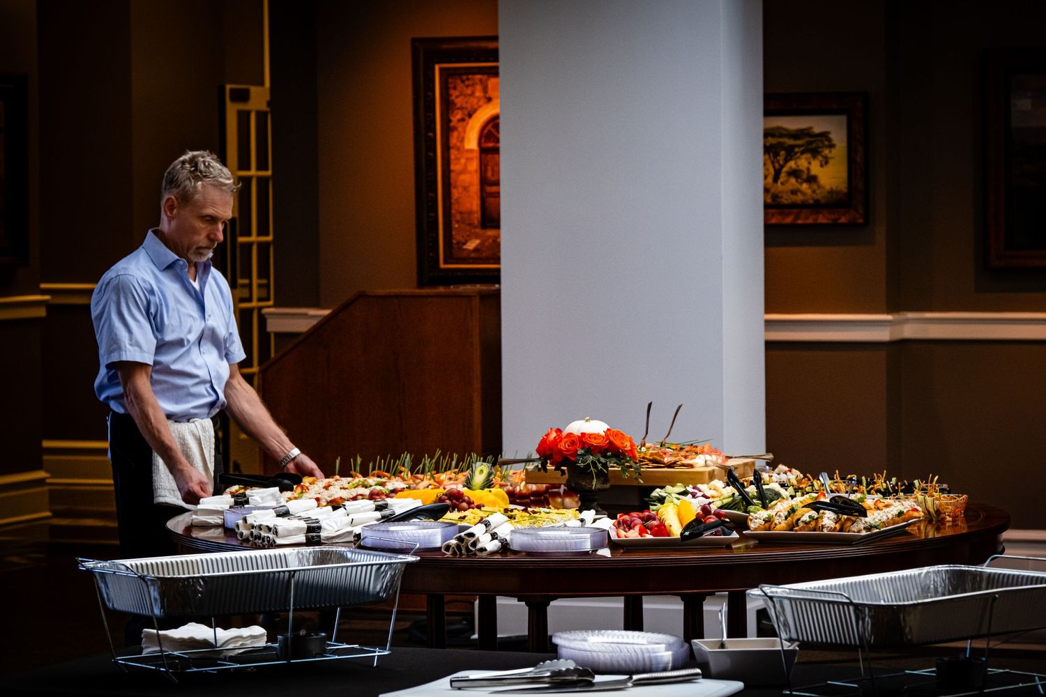 Man carefully arranging an extensive catered food spread on a large table in an elegant venue.