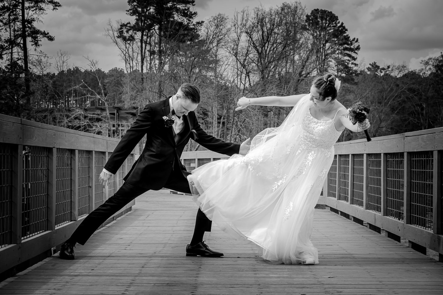 Black and white photo of bride and groom playfully dancing on a wooden bridge on their wedding day.