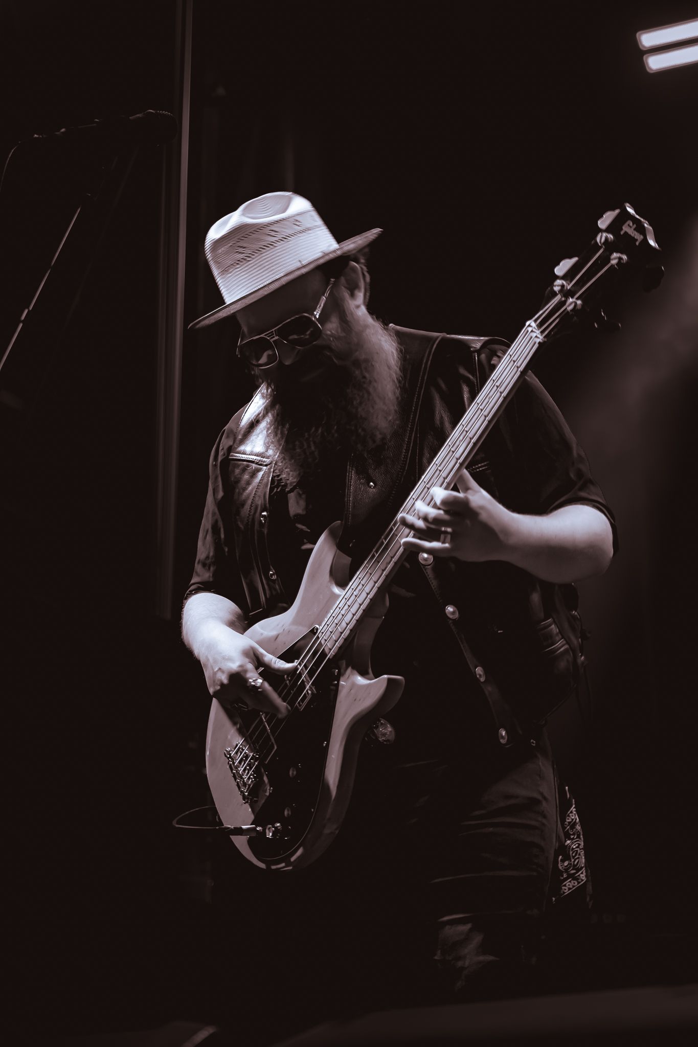 Black and white photo of a bearded male bassist wearing a hat and sunglasses performing on stage.