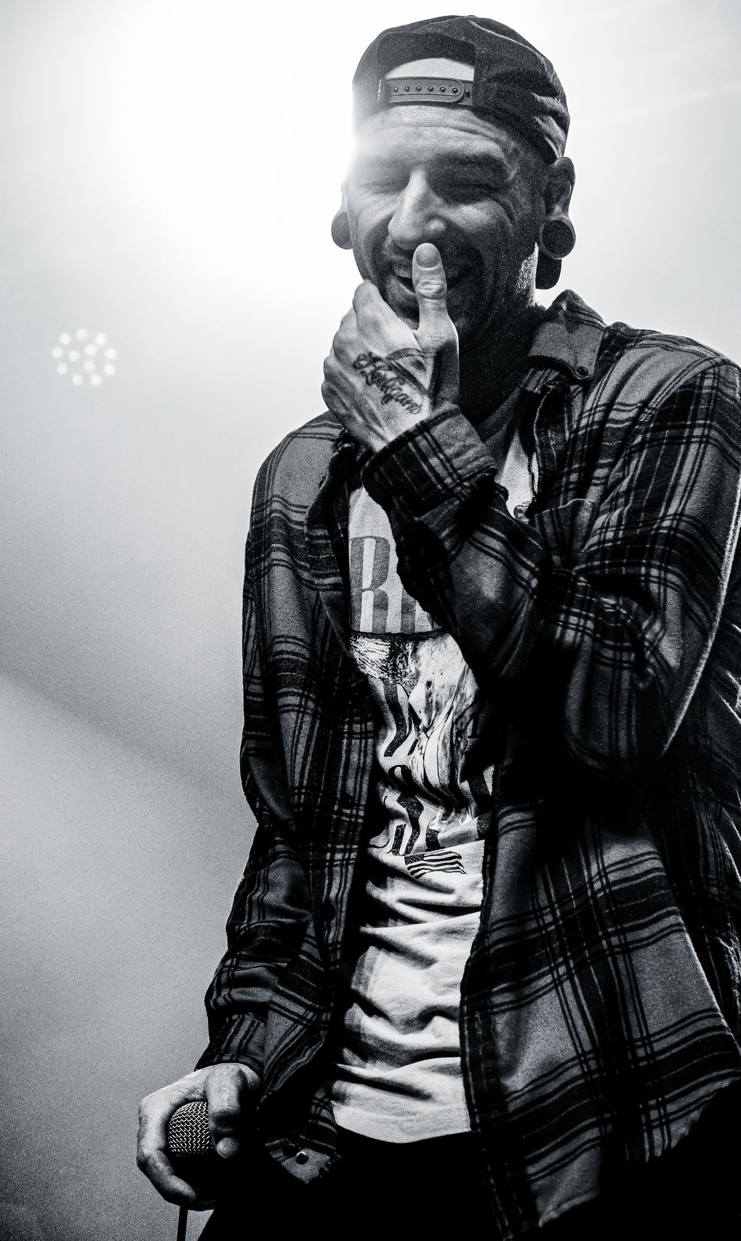 Black and white photo of a smiling male vocalist holding a microphone, wearing a plaid shirt and snapback cap.
