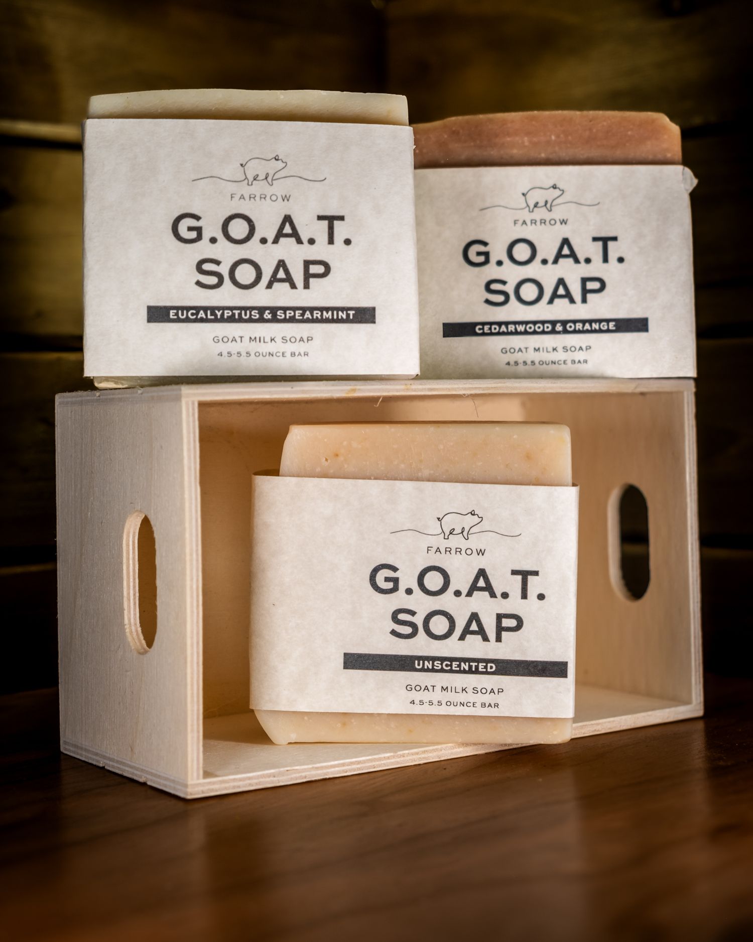 Three bars of G.O.A.T. goat milk soap in kraft paper packaging displayed in a wooden crate.  Product photography. Branding materials. Brands