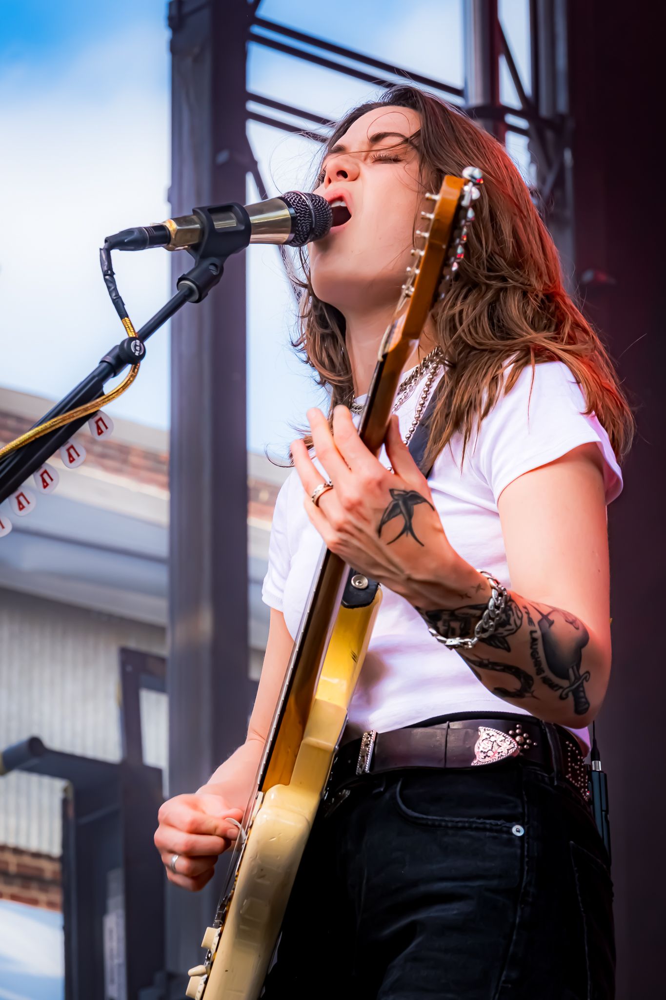 Tattooed female vocalist playing gold guitar and singing at outdoor stage in white tee.