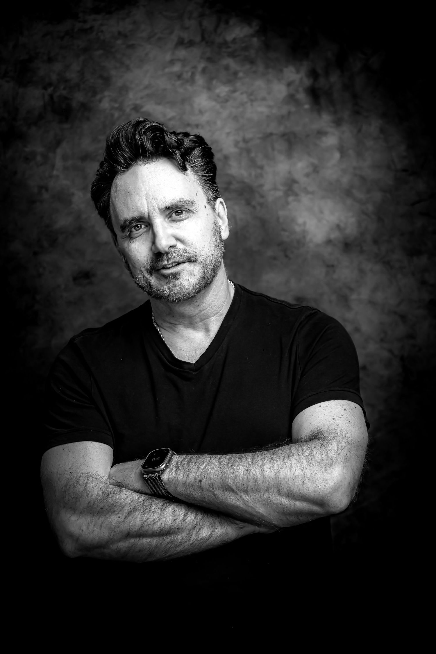 Black and white portrait of a middle-aged man in a black t-shirt, arms crossed, wearing a watch.