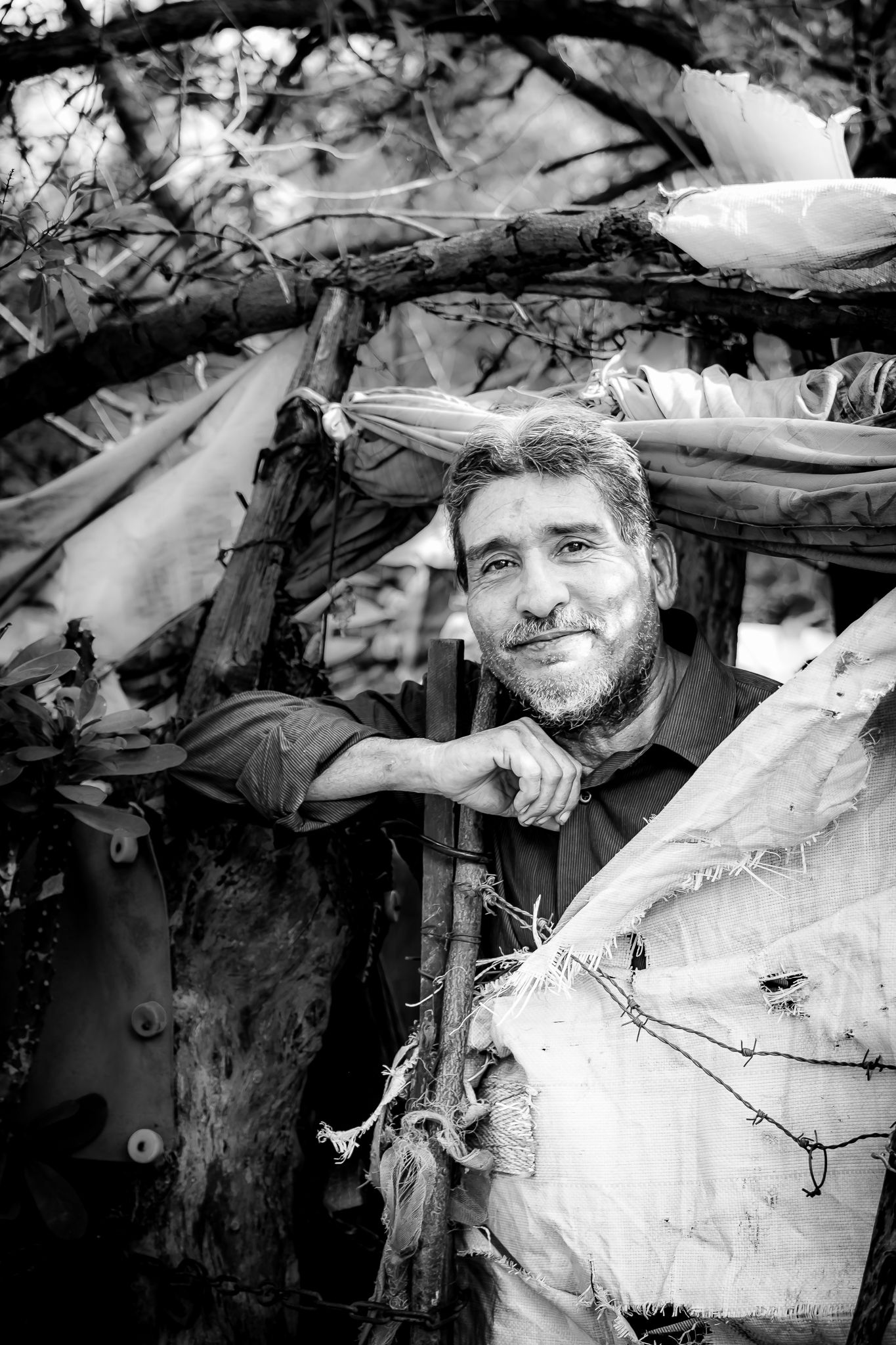 Smiling bearded man leaning on wooden post surrounded by rustic materials in black and white.