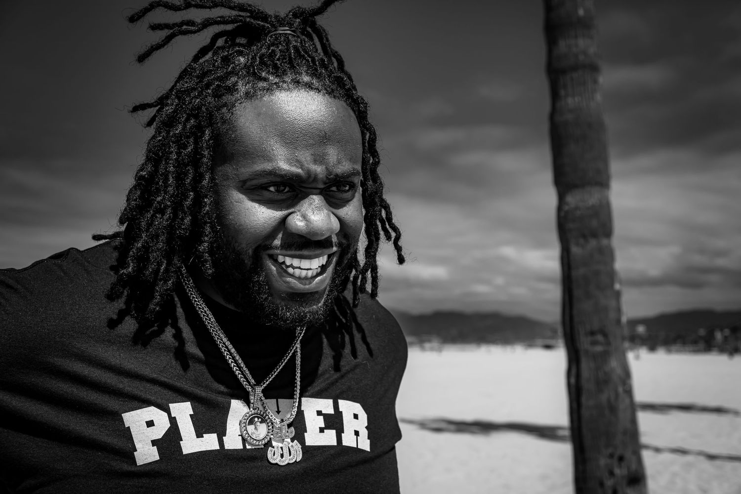 Laughing man with dreadlocks wearing PLAYER t-shirt enjoys a moment on a beach in black and white photo.