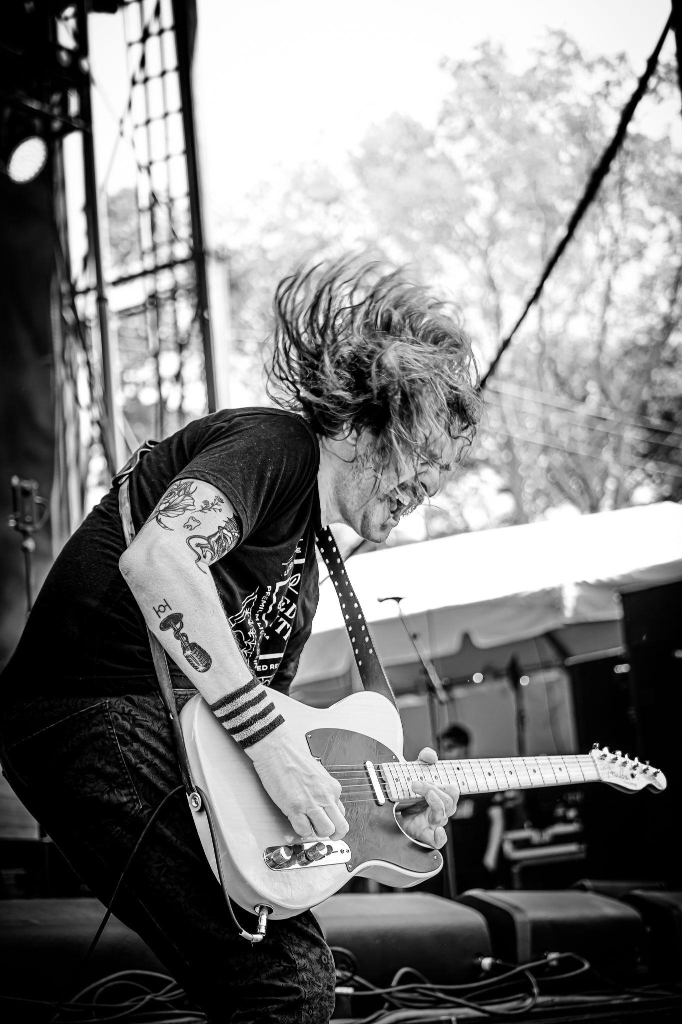 Tattooed guitarist headbanging while playing electric guitar on outdoor stage in black and white.