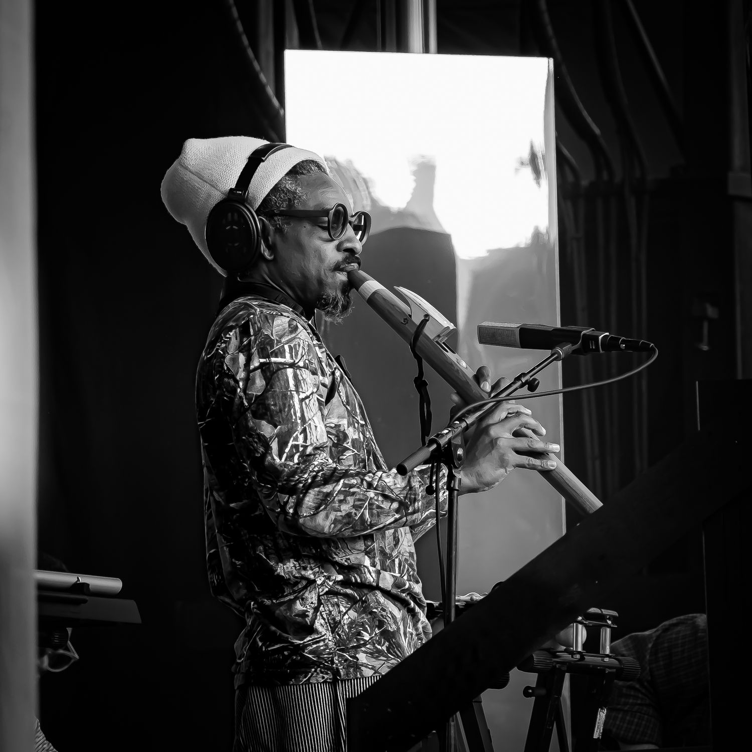 Black and white photo of a musician playing a wind instrument at a microphone, wearing headphones and a patterned shirt.