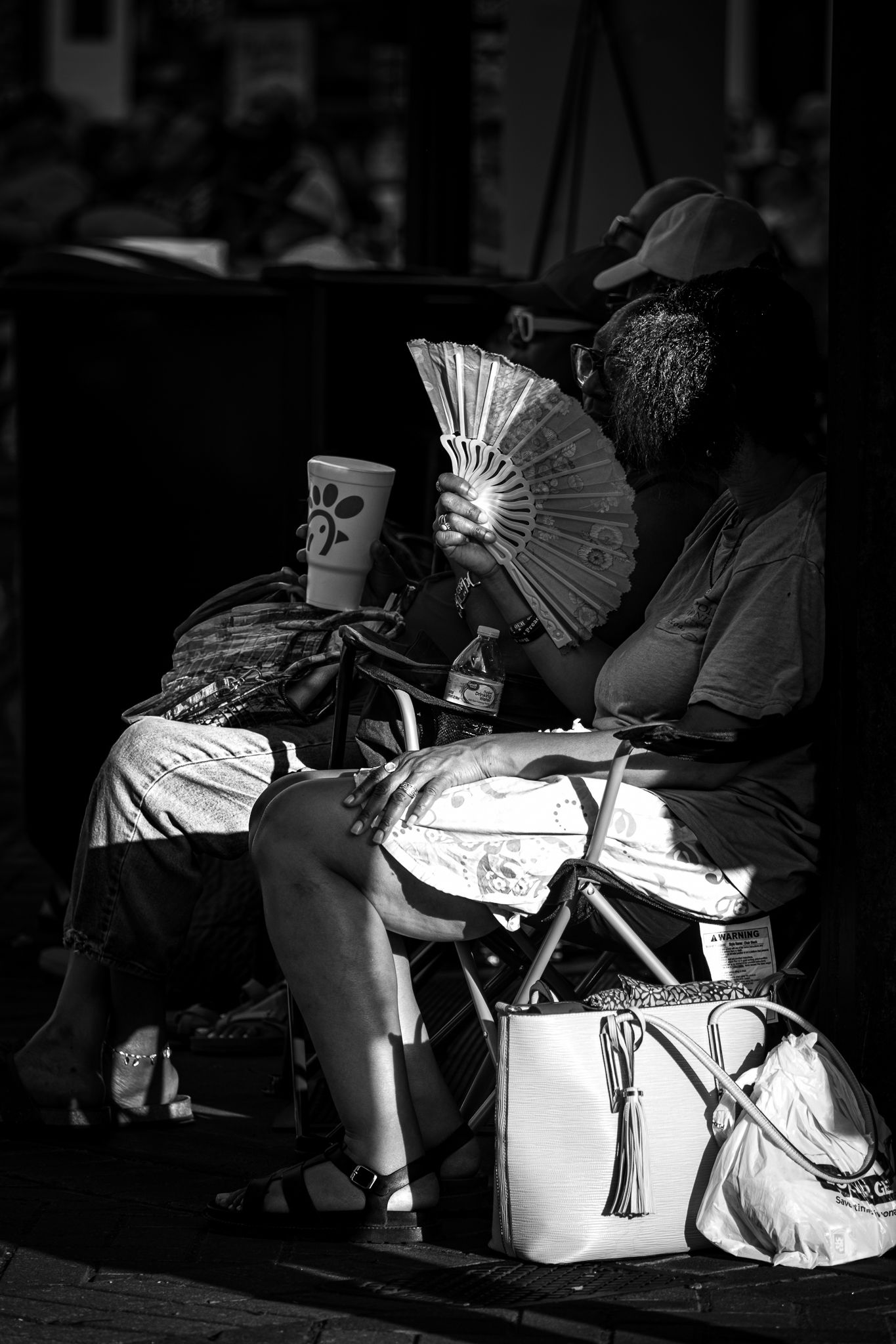 Woman sitting in chair holding decorative fan and Chick-fil-A cup, black and white street photography.