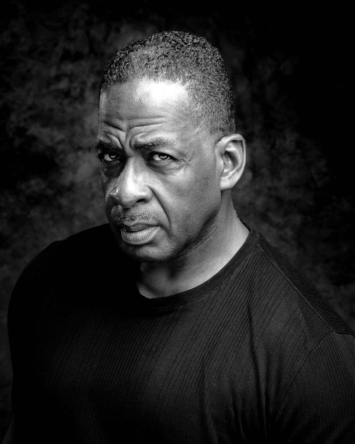 Black and white portrait of a serious middle-aged man in a black shirt against a dark background.