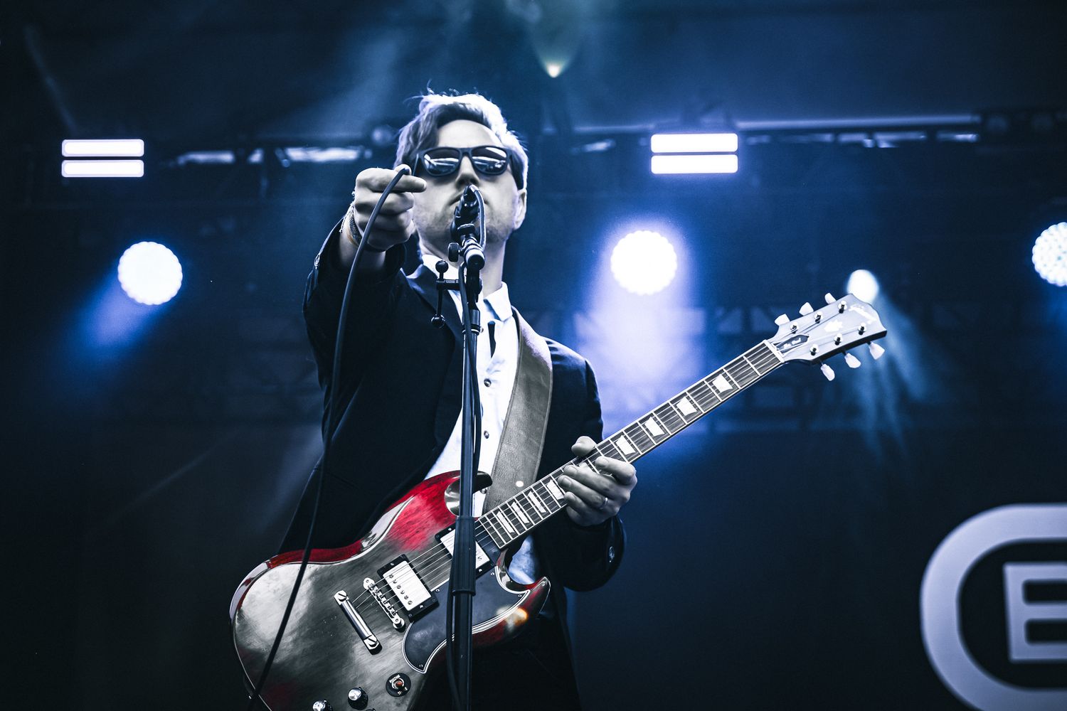 Male guitarist in suit pointing at crowd while performing with red electric guitar on stage.