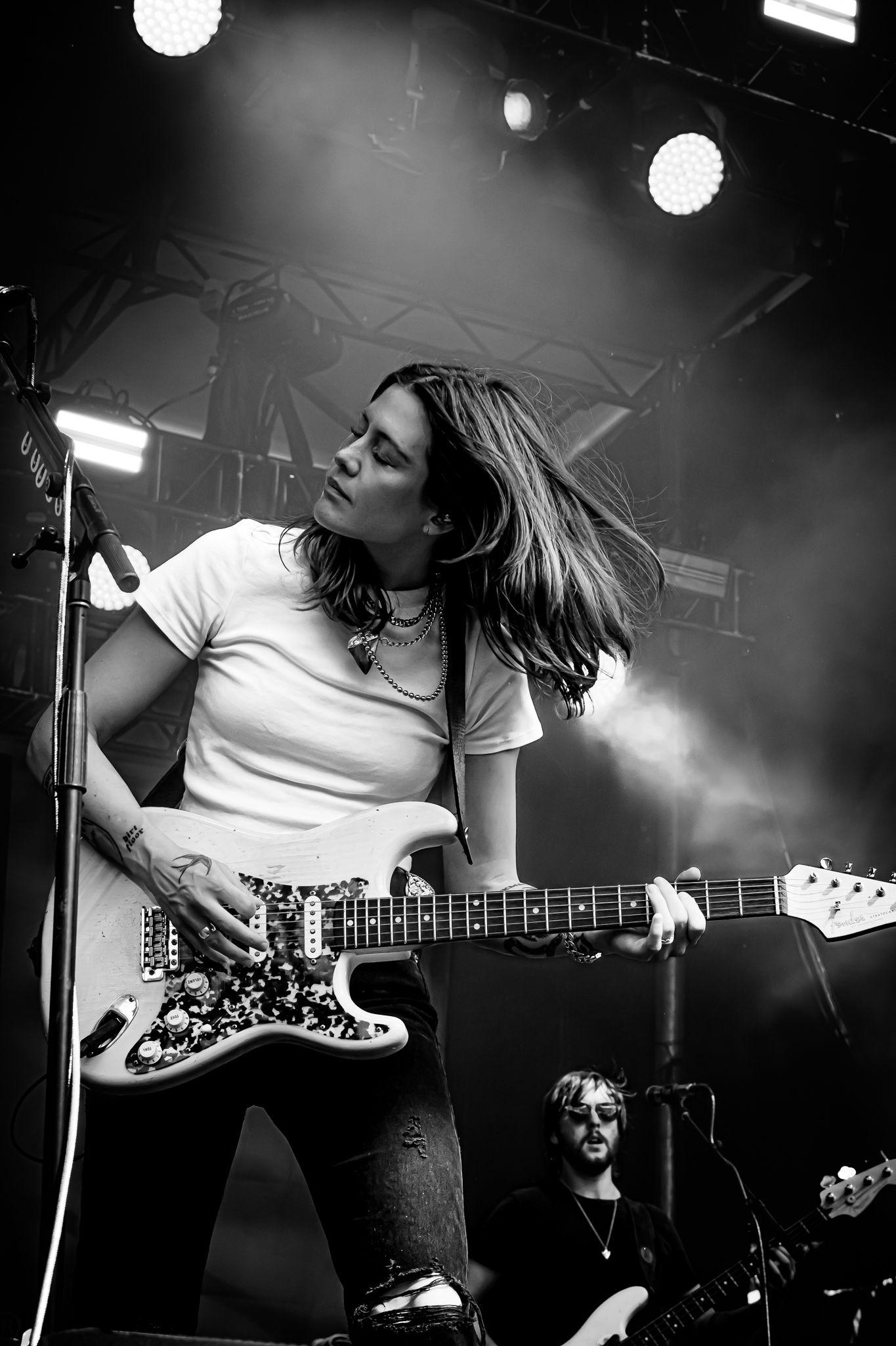 Female guitarist tossing hair while playing Fender guitar on dramatic smoky stage in black and white.