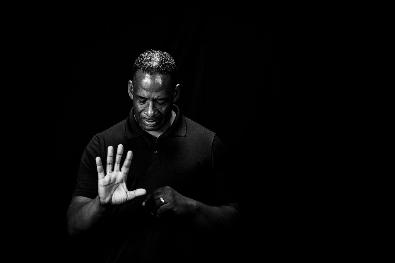 Black and white portrait of a man in a black polo raising his hand against a dark background.