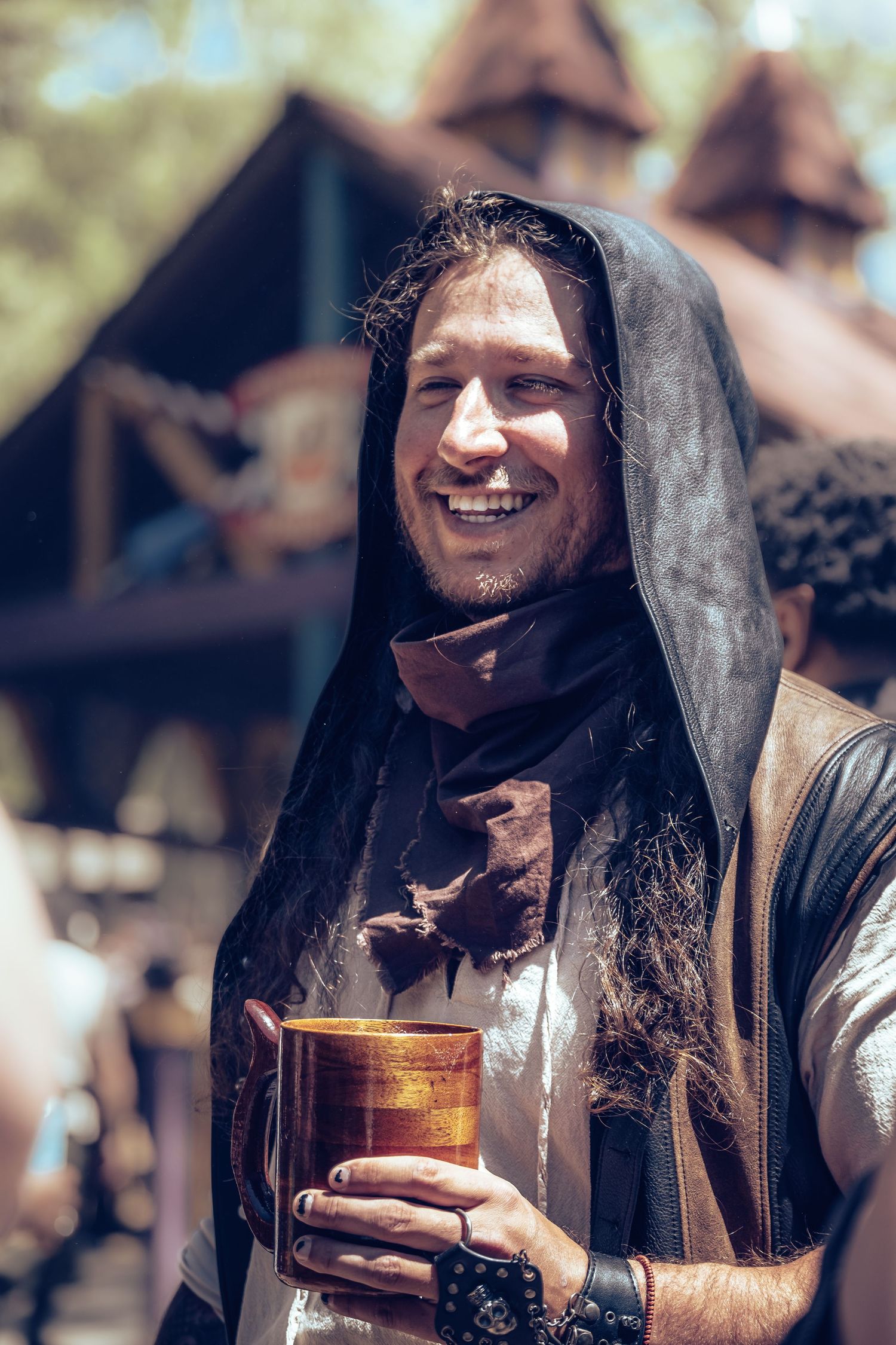 Smiling man with long hair wearing a dark hooded cloak holds a copper mug at a Renaissance faire.