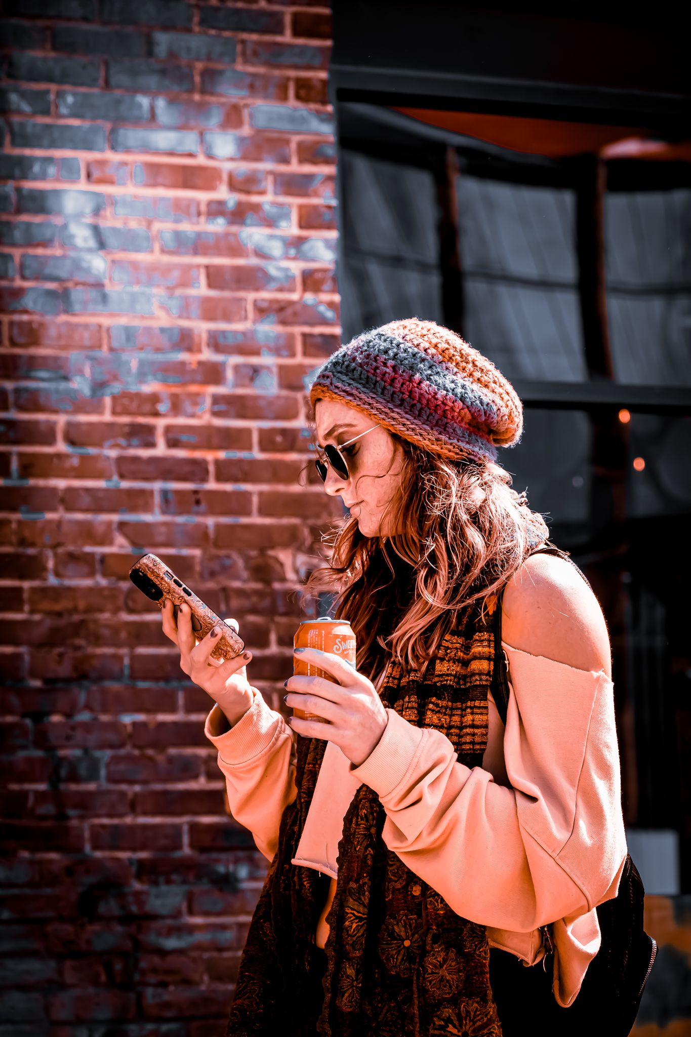 Stylish woman in a knit beret and sunglasses checks her phone while holding a drink near a brick wall outdoors.