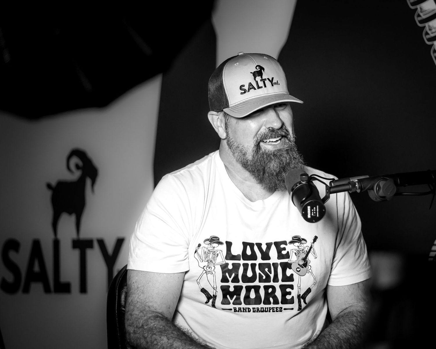 Bearded man in Salty cap laughs at microphone in black and white music studio portrait. Podcast coverage. Branding. Branding material.