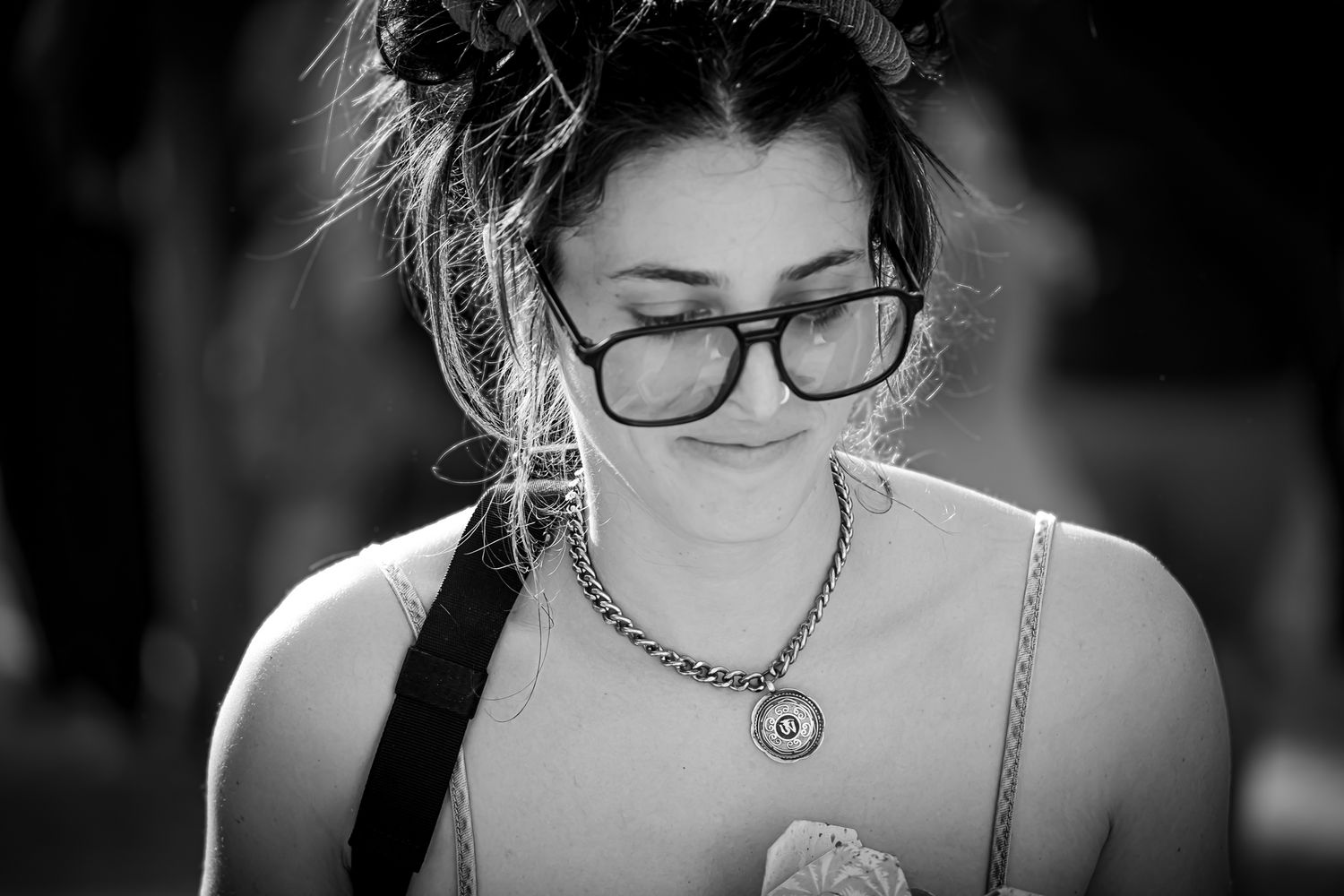 Black and white portrait of a young woman wearing thick-rimmed glasses and a chain necklace, looking down.