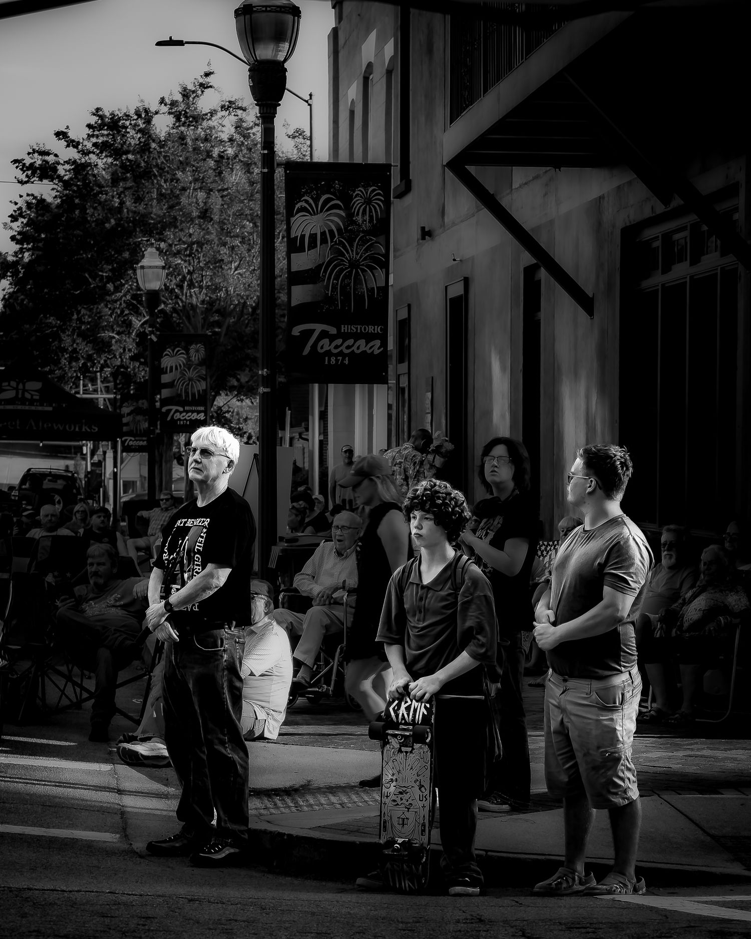 Black and white street photo of people standing on a sidewalk near a lamp post outside the Toccoa cafe.