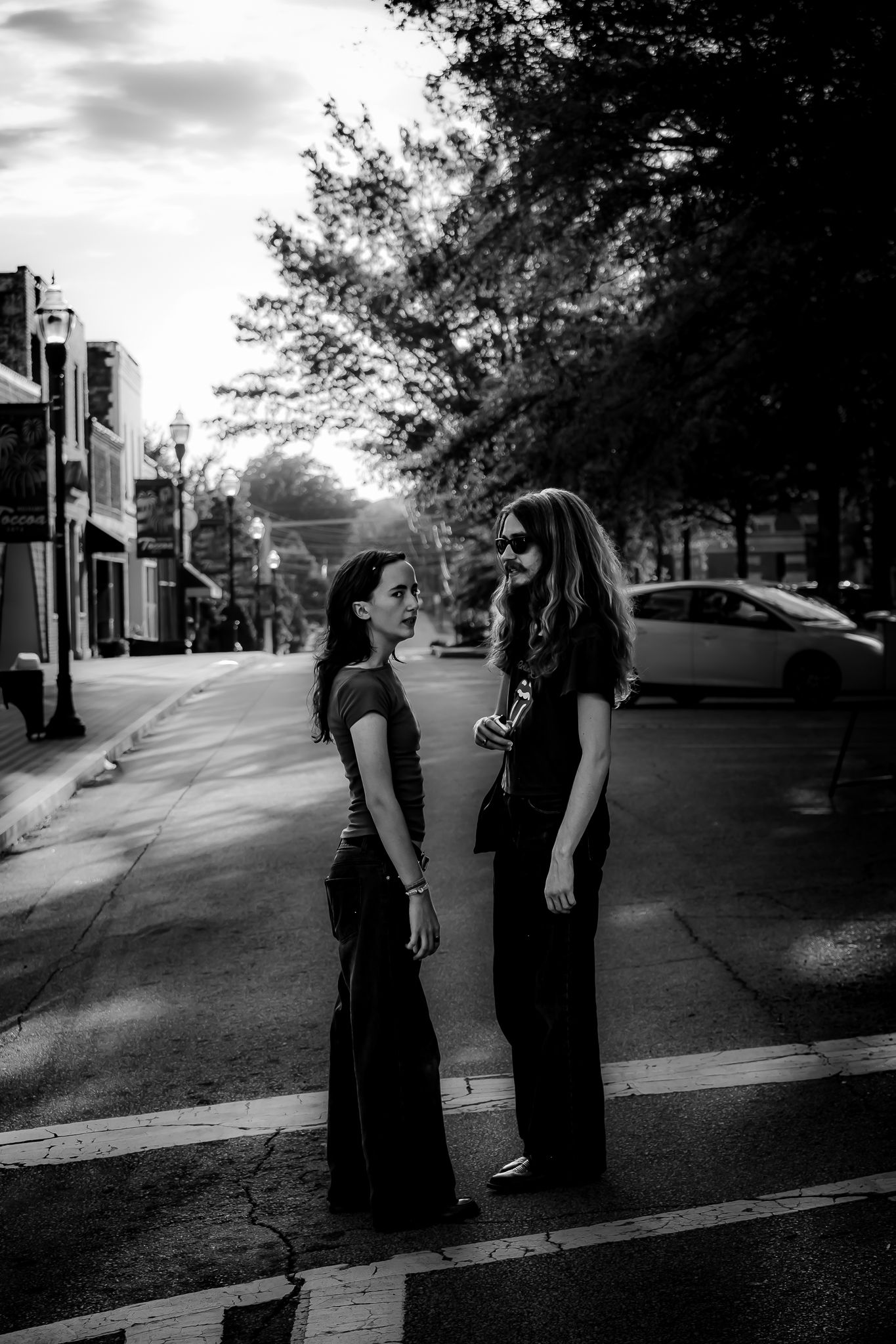 Two women stand facing each other on a city street in black and white photo with trees and parked cars in background.