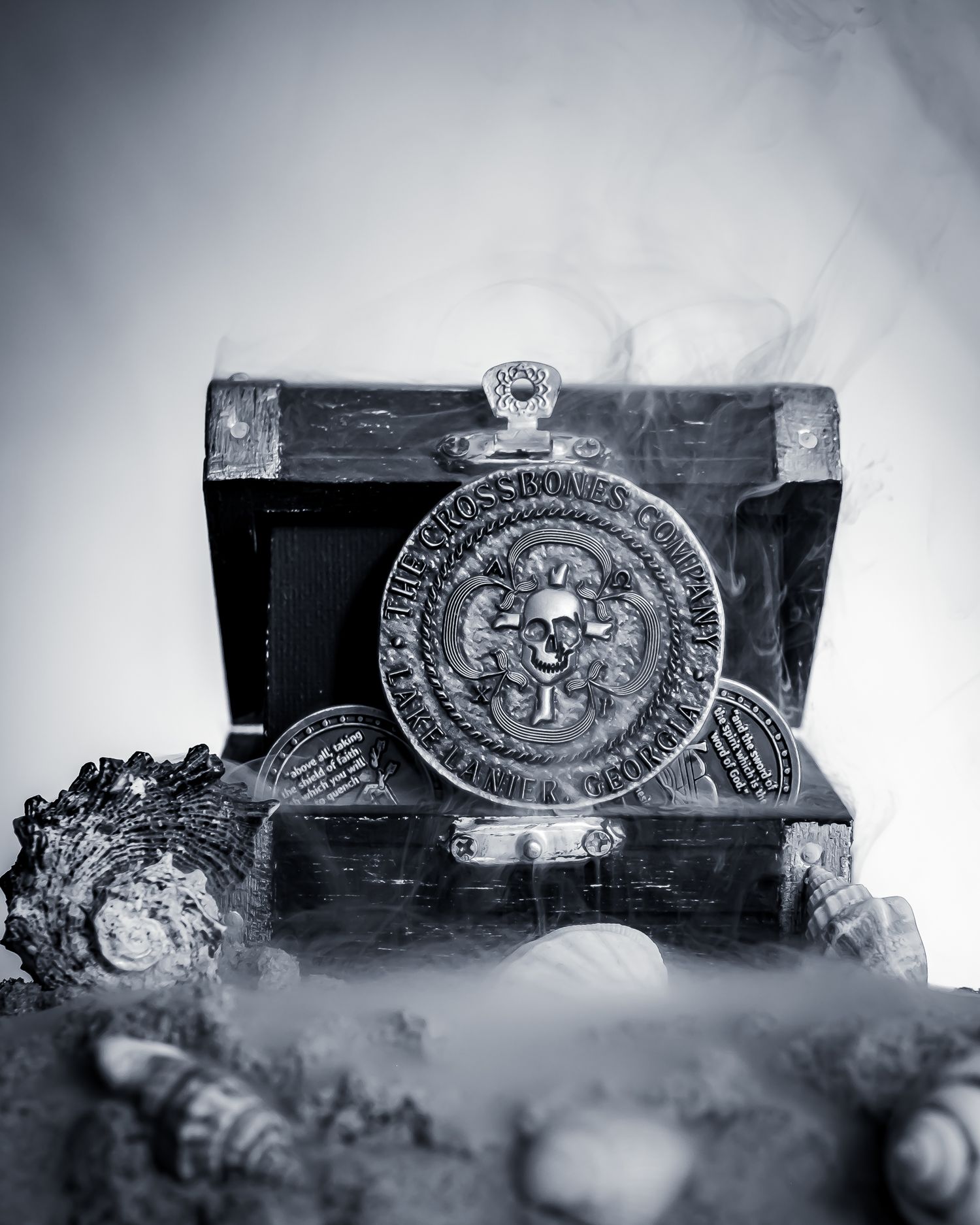 Black and white photo of a pirate treasure chest with skull coins and seashells surrounded by mystical smoke. Branding materials. product.